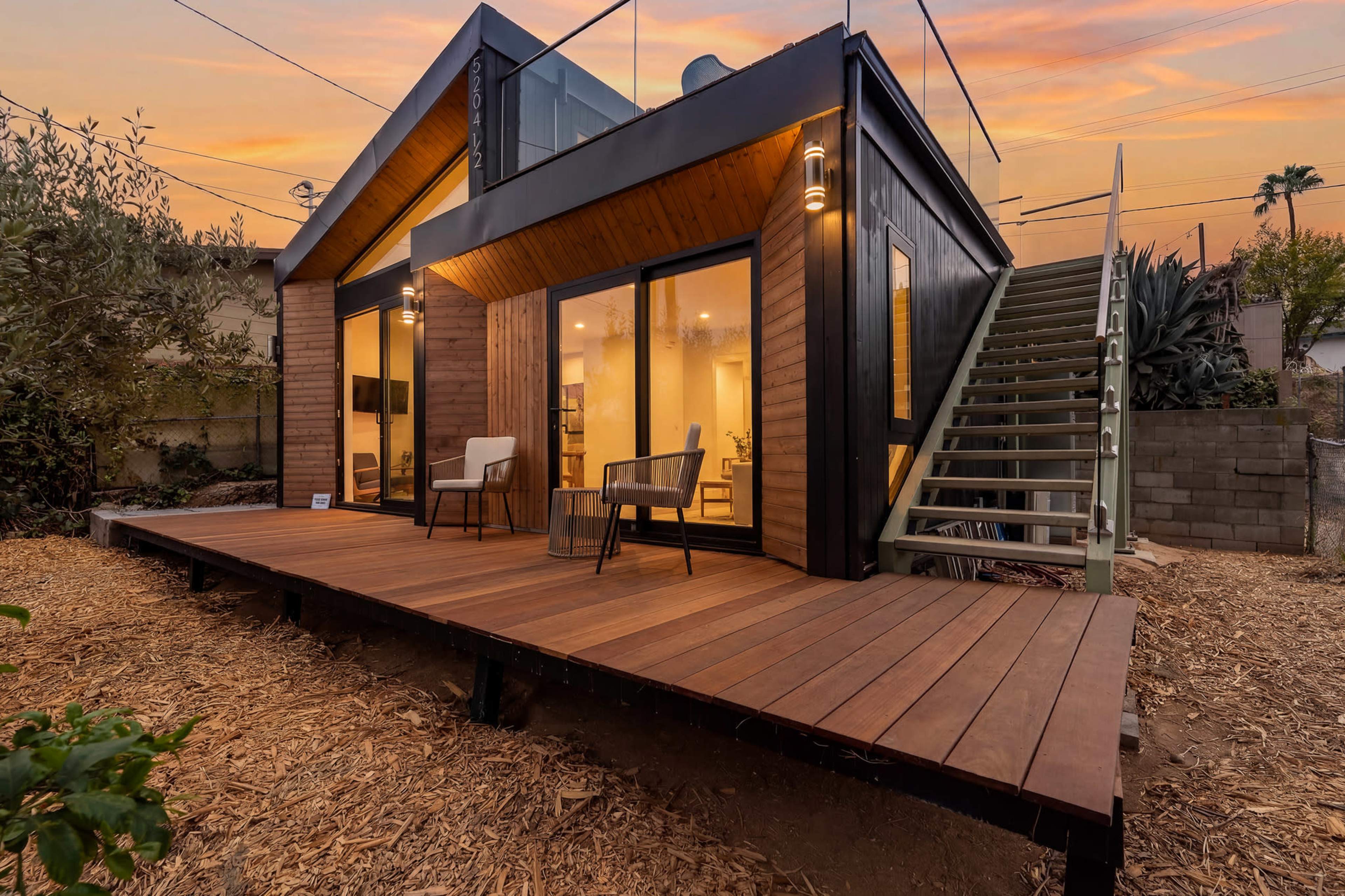 A modern cabin features a wooden deck, large windows, and a staircase leading to an upper level, set against a colorful sunset sky.