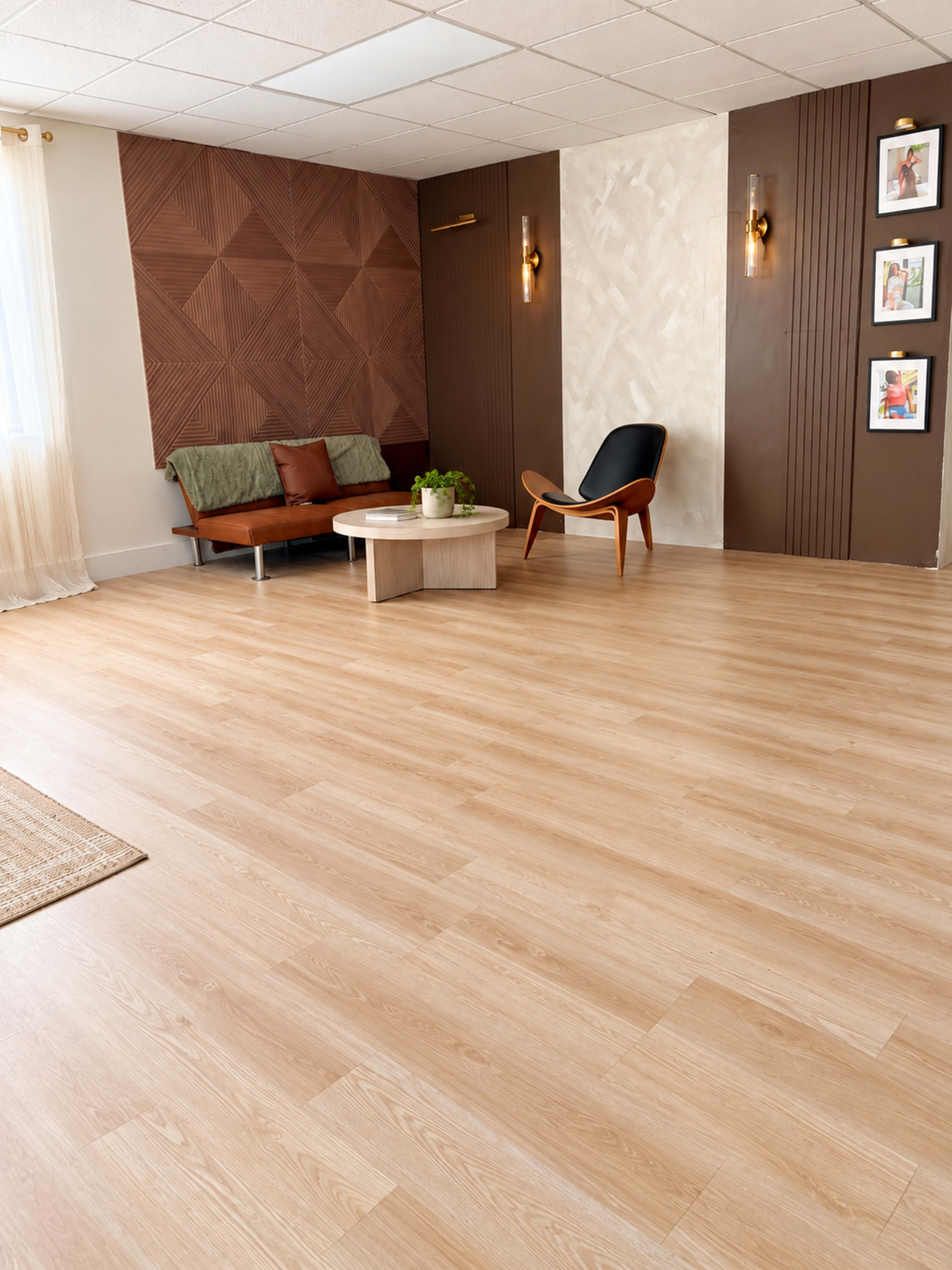 The image features a spacious and well-lit interior with wooden flooring, a brown accent wall with geometric decor, a green couch, a round coffee table, and framed pictures on the wall.