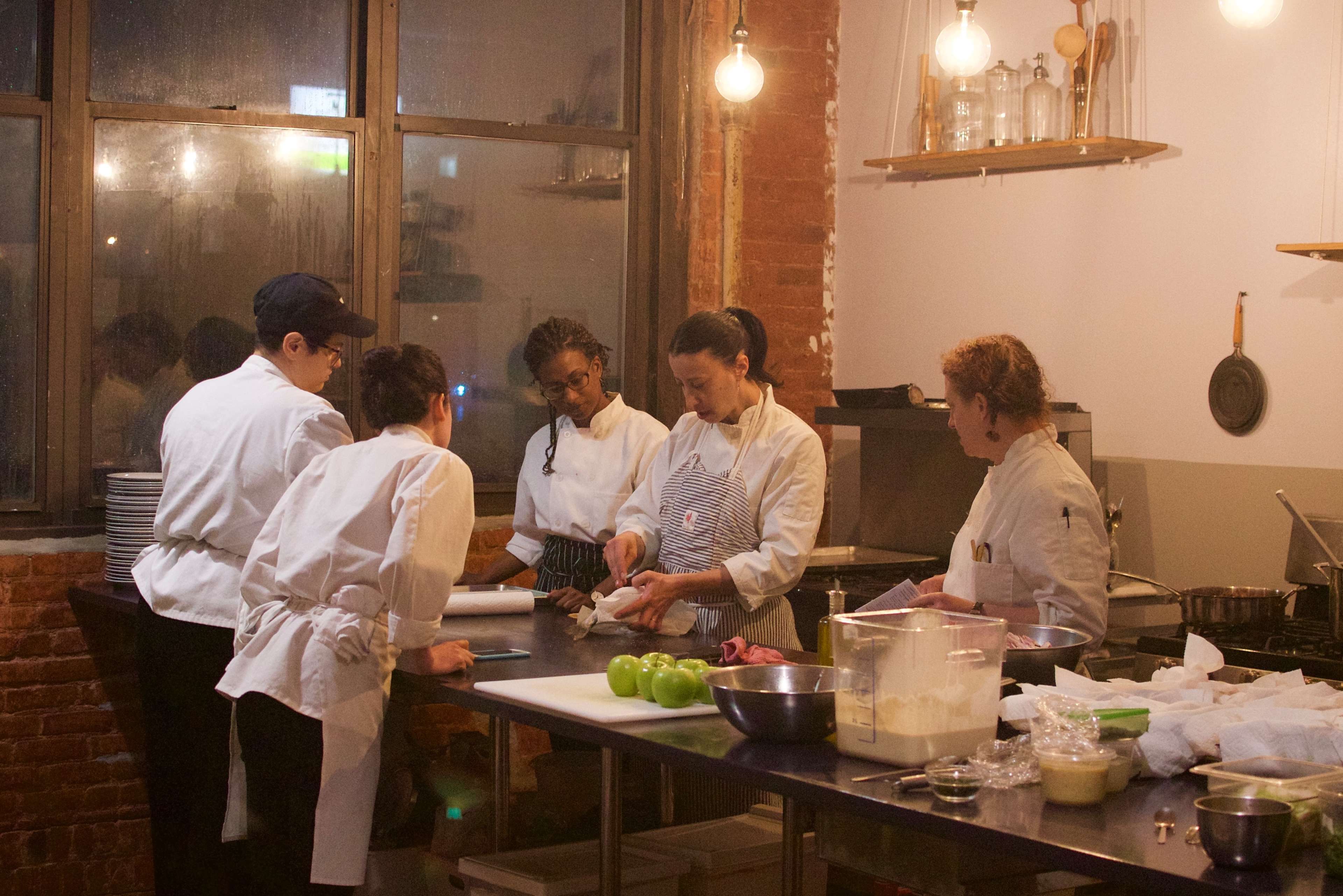 A group of five chefs in white uniforms prepares food at a kitchen counter illuminated by hanging lights.