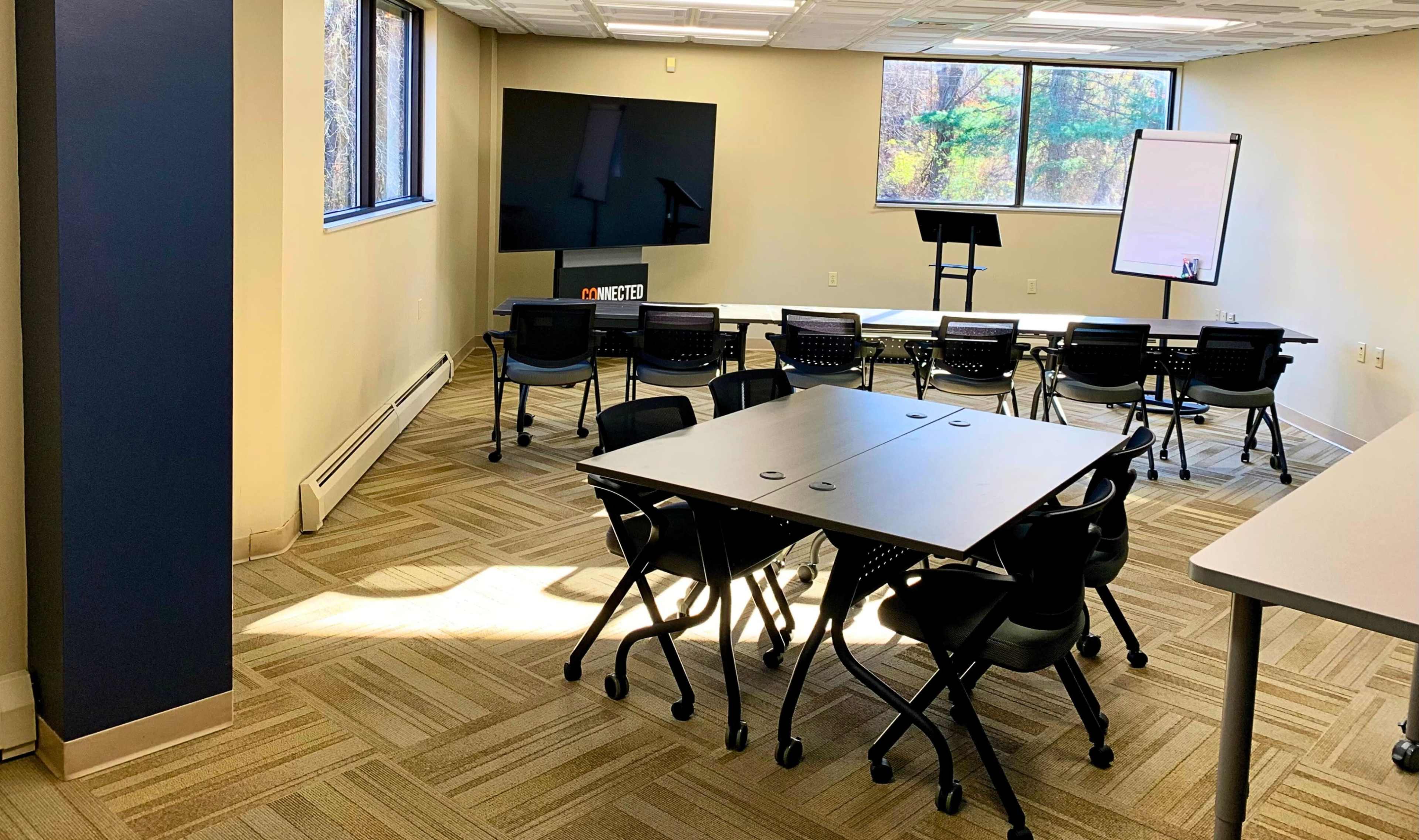 The image shows a light-filled conference room with several tables and chairs arranged for a meeting, featuring a large screen and a whiteboard along the walls.