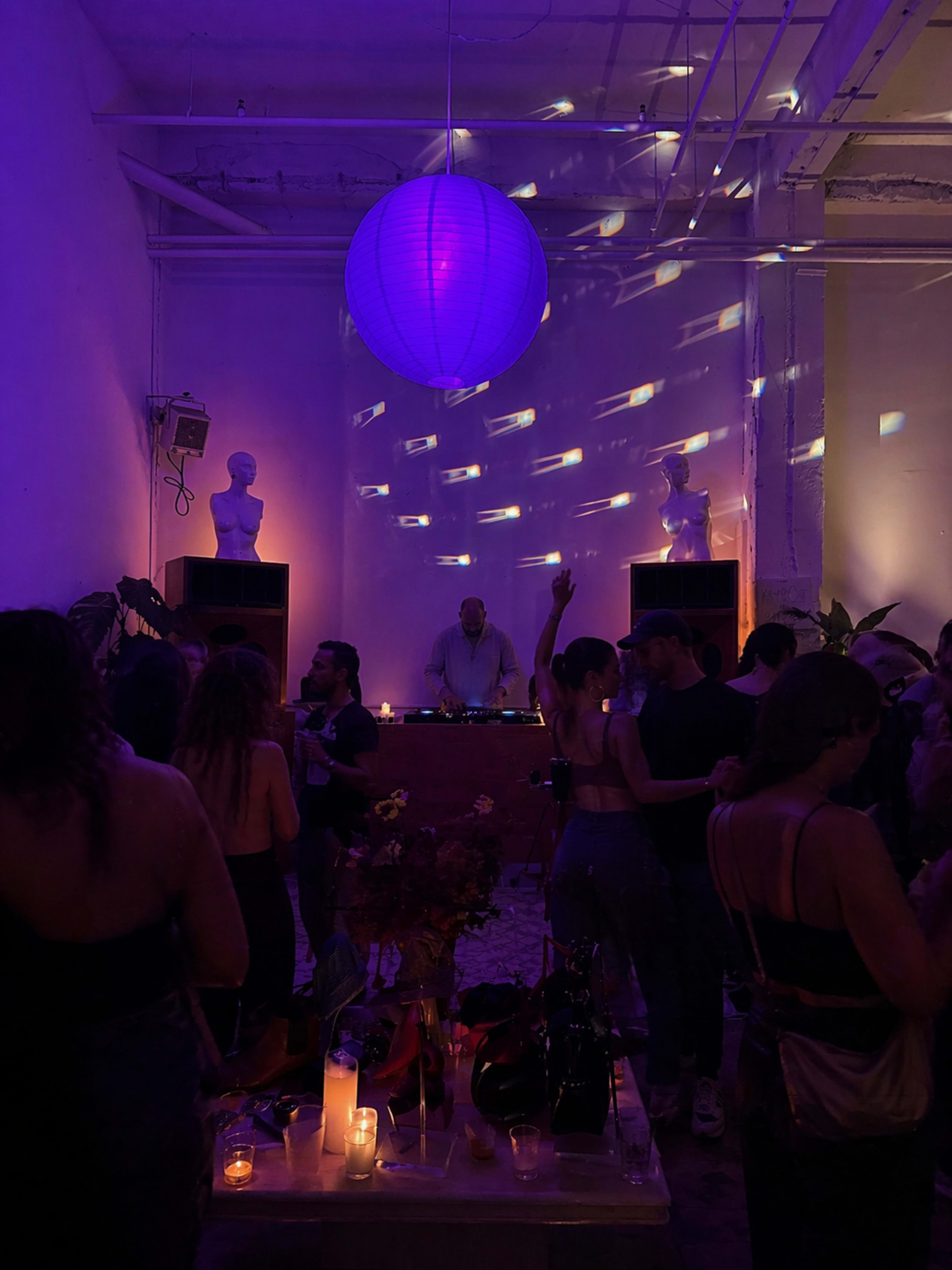A DJ plays music in a dimly lit venue filled with people dancing, surrounded by purple lighting and decorative elements.