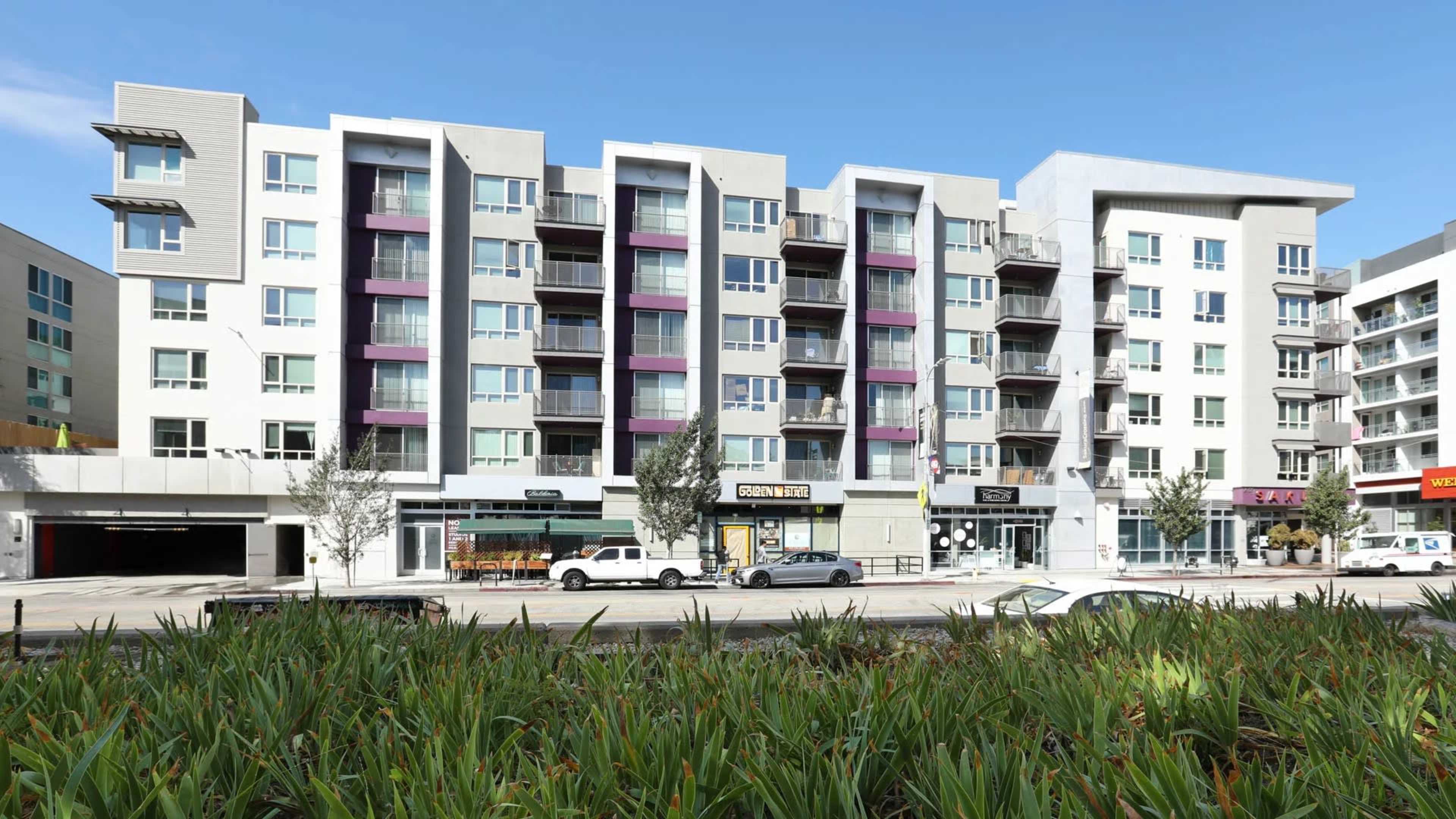The image shows a medium-sized apartment building with multiple balconies and ground-level commercial spaces, surrounded by landscaped greenery.
