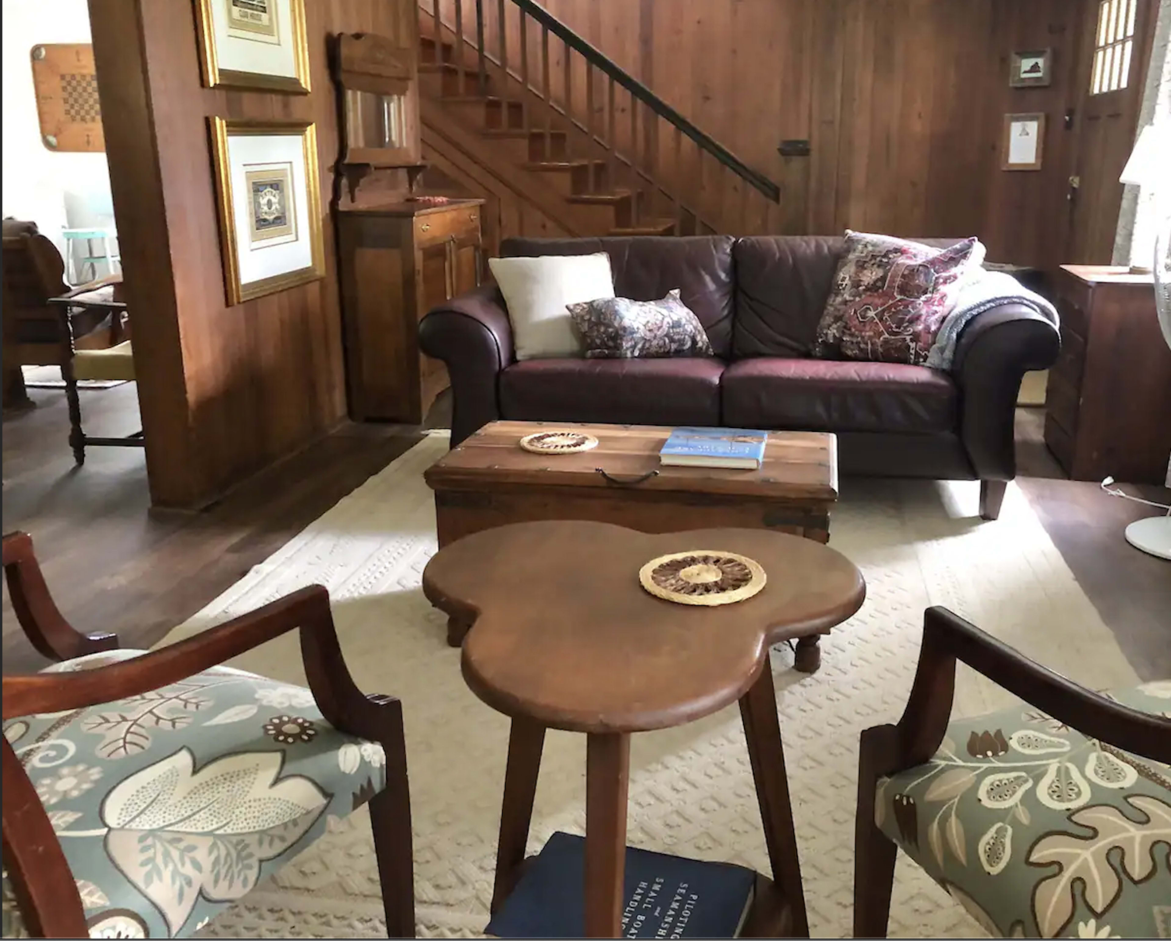 The scene features a living room with a brown leather sofa, a wooden coffee table, and two patterned chairs, all set against a backdrop of wooden walls and a staircase.