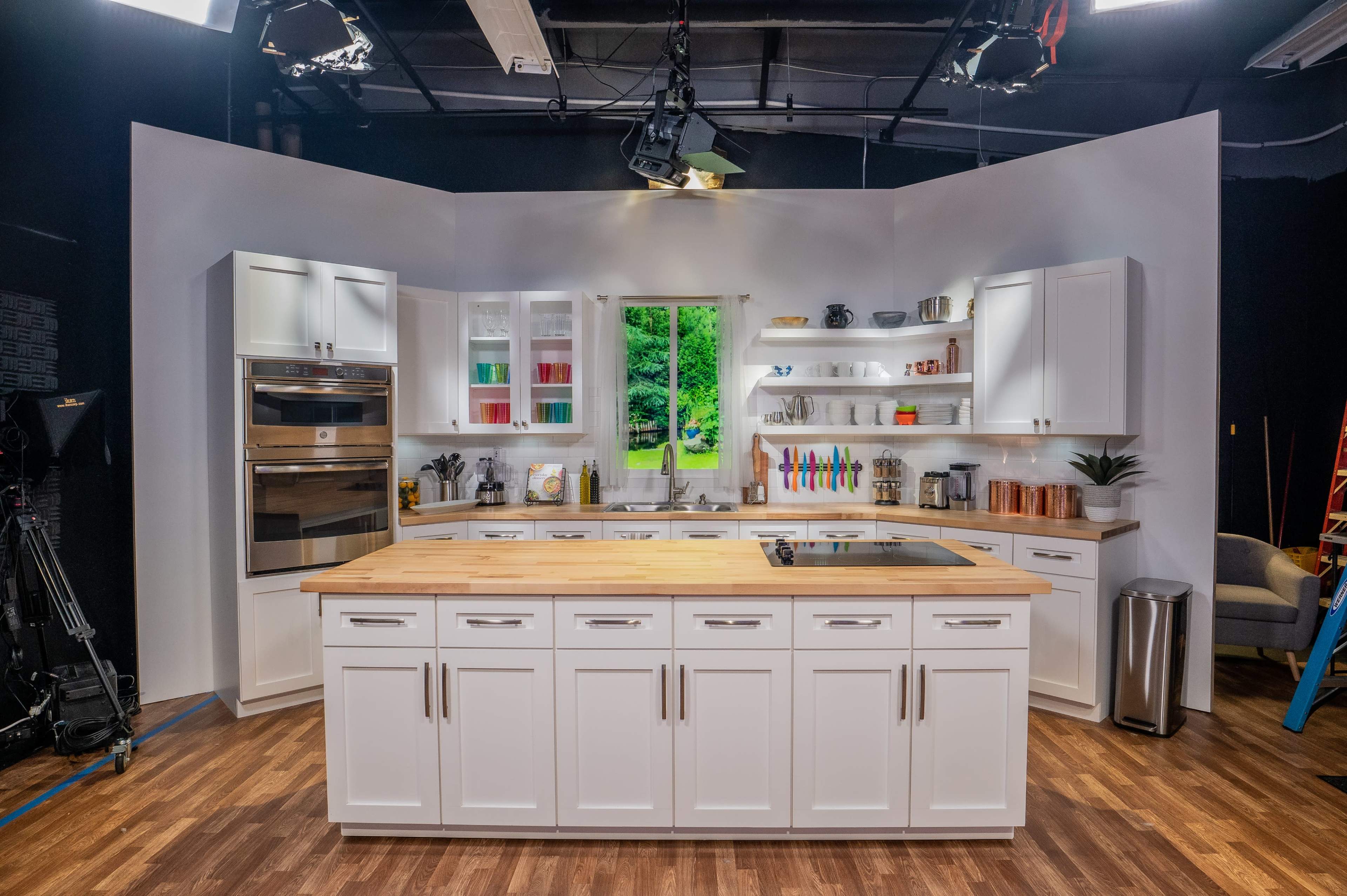 A modern kitchen set features a central island with a wooden countertop, surrounded by white cabinetry and shelves filled with kitchenware, set against a backdrop of a large window offering a view of greenery.
