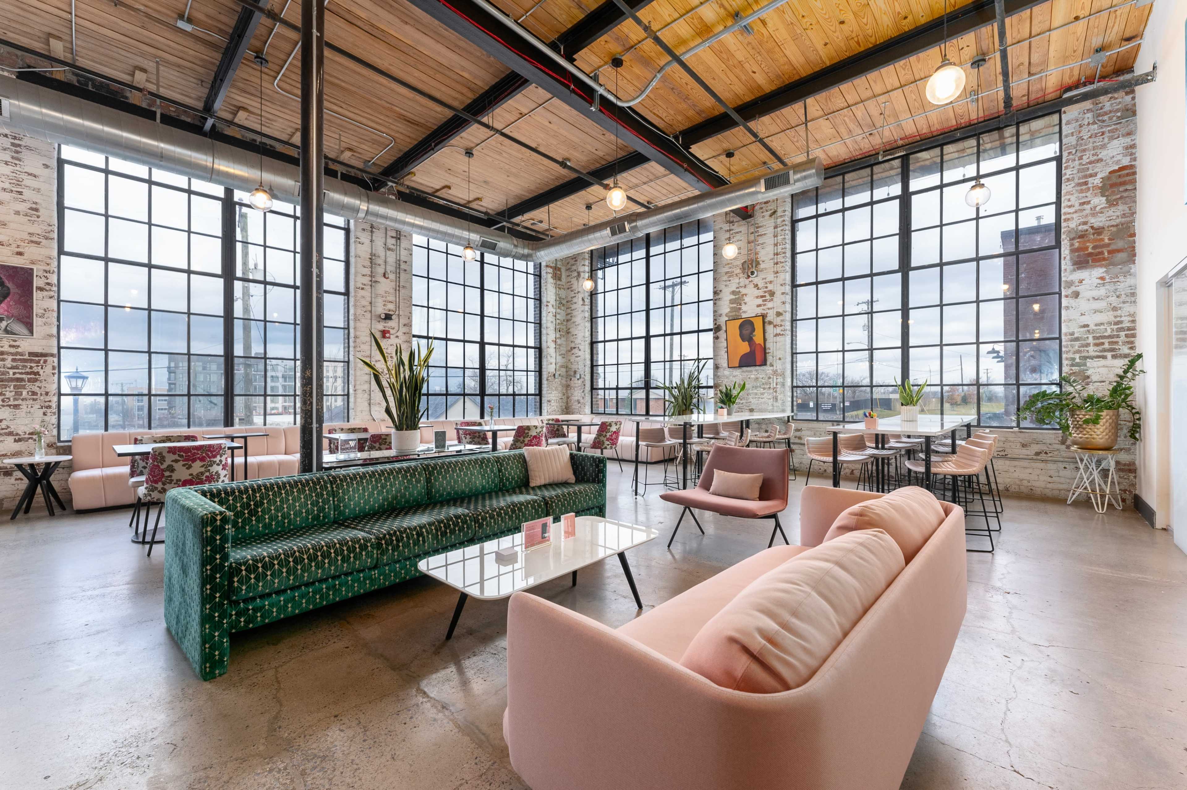 A modern café with large windows, exposed brick walls, and a mix of green and pink upholstered seating arrangements.
