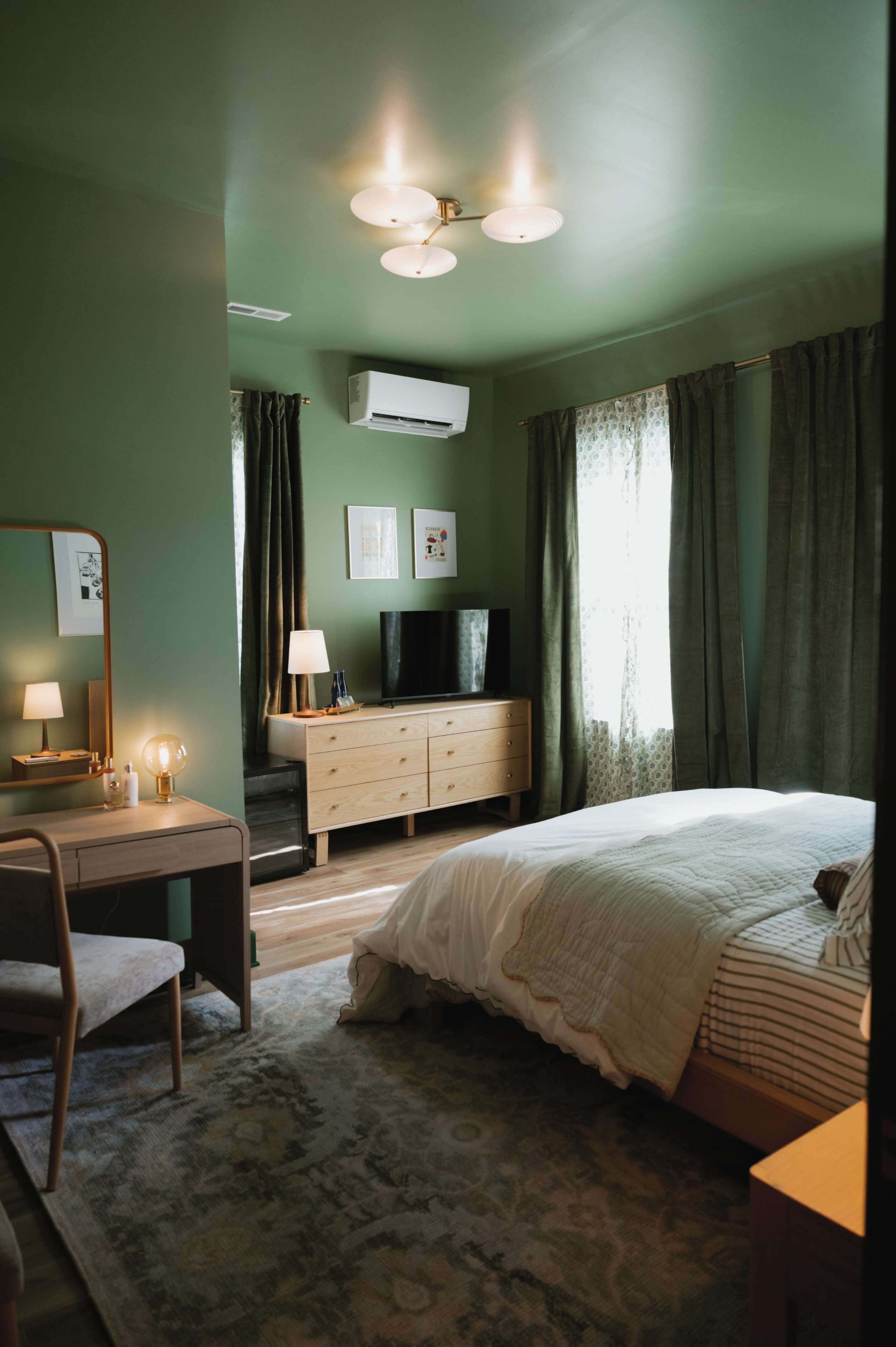 The image shows a well-decorated bedroom with green walls, a bed with a light-colored quilt, a TV on a wooden dresser, and a chair next to a vanity table.