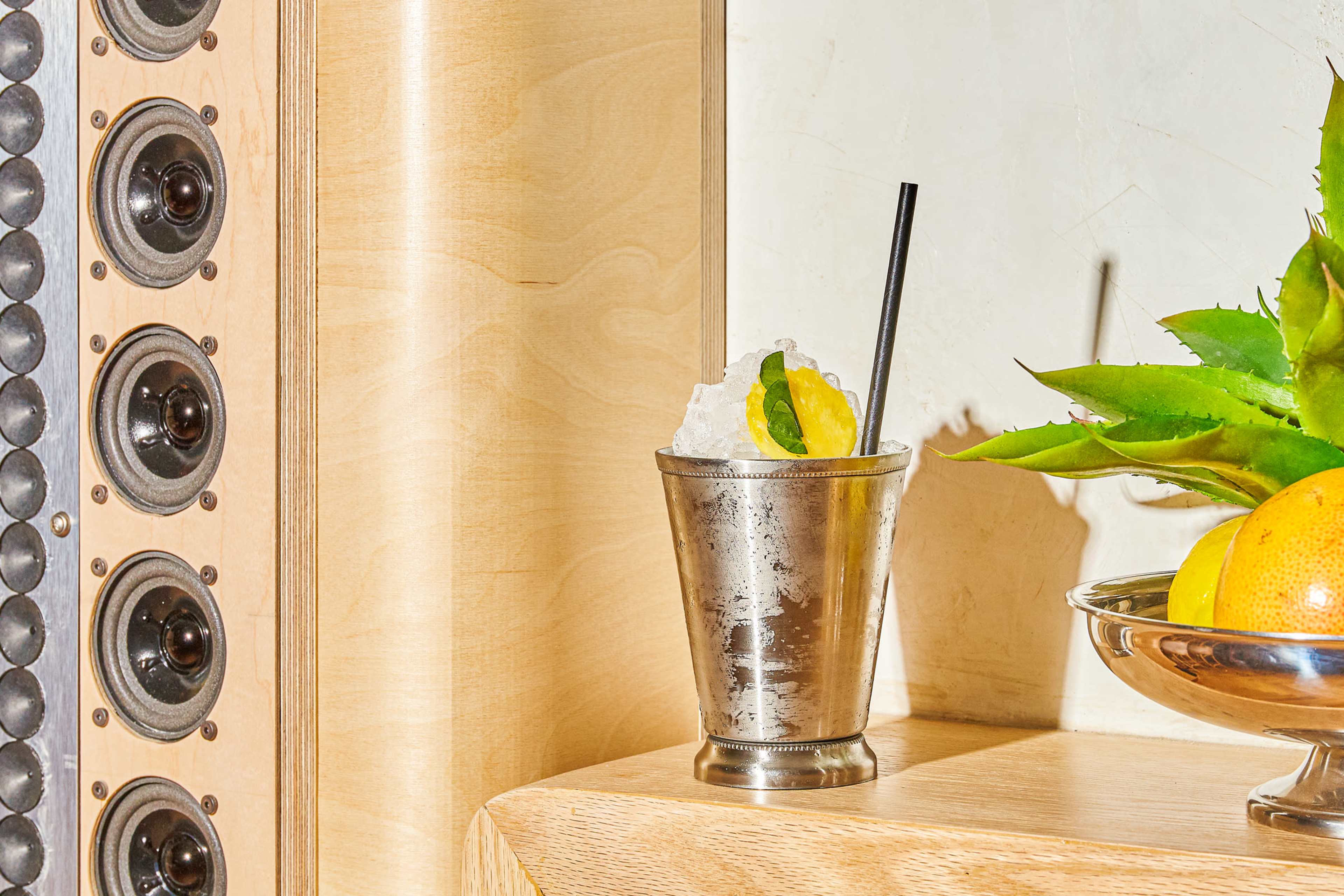 A silver cocktail glass filled with crushed ice, a lime wedge, and a black straw sits next to a bowl of oranges on a wooden surface, with speaker grills in the background.