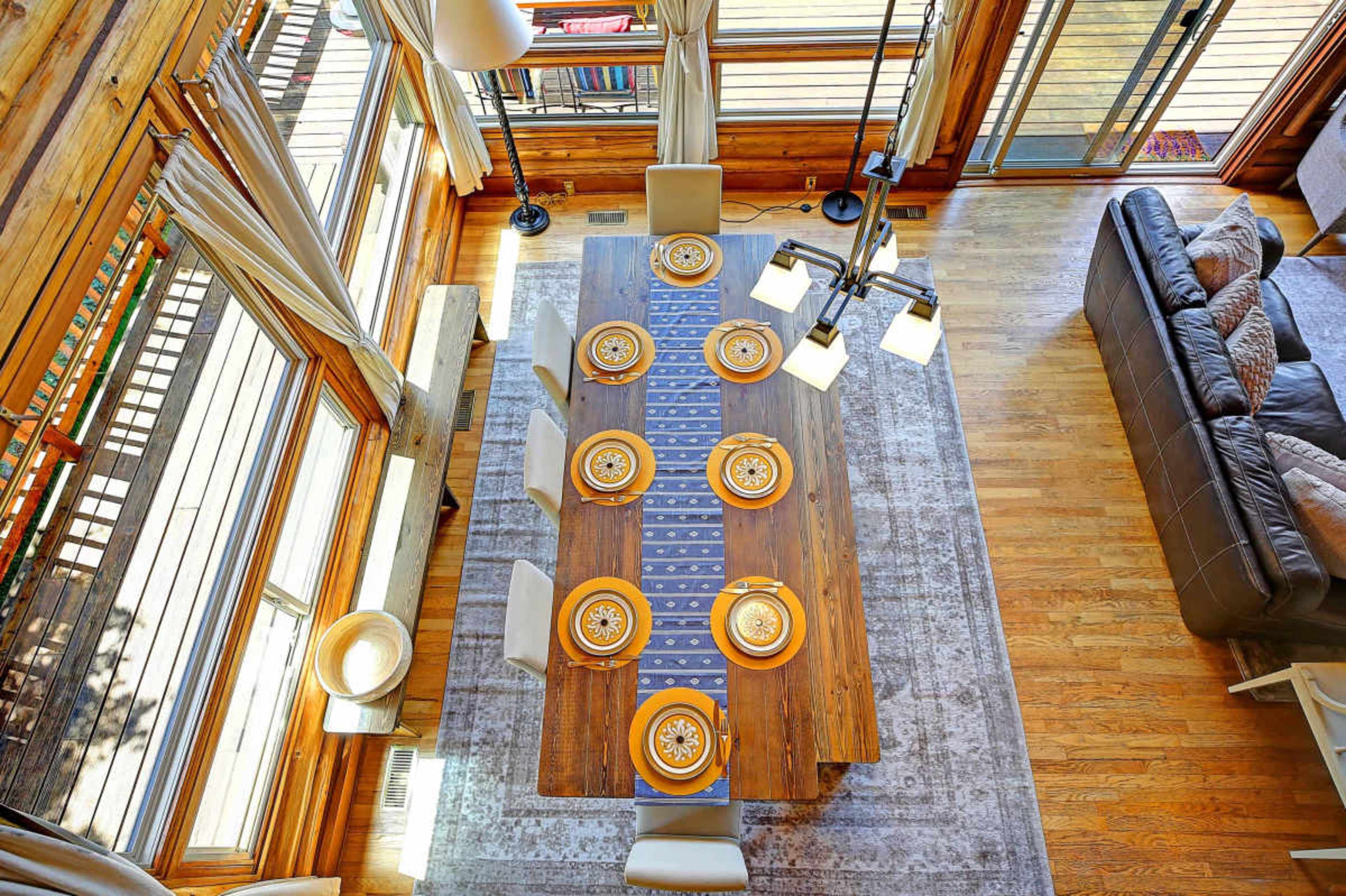 A wooden dining table is set with place settings and a decorative runner in a brightly lit room with large windows and a seating area nearby.