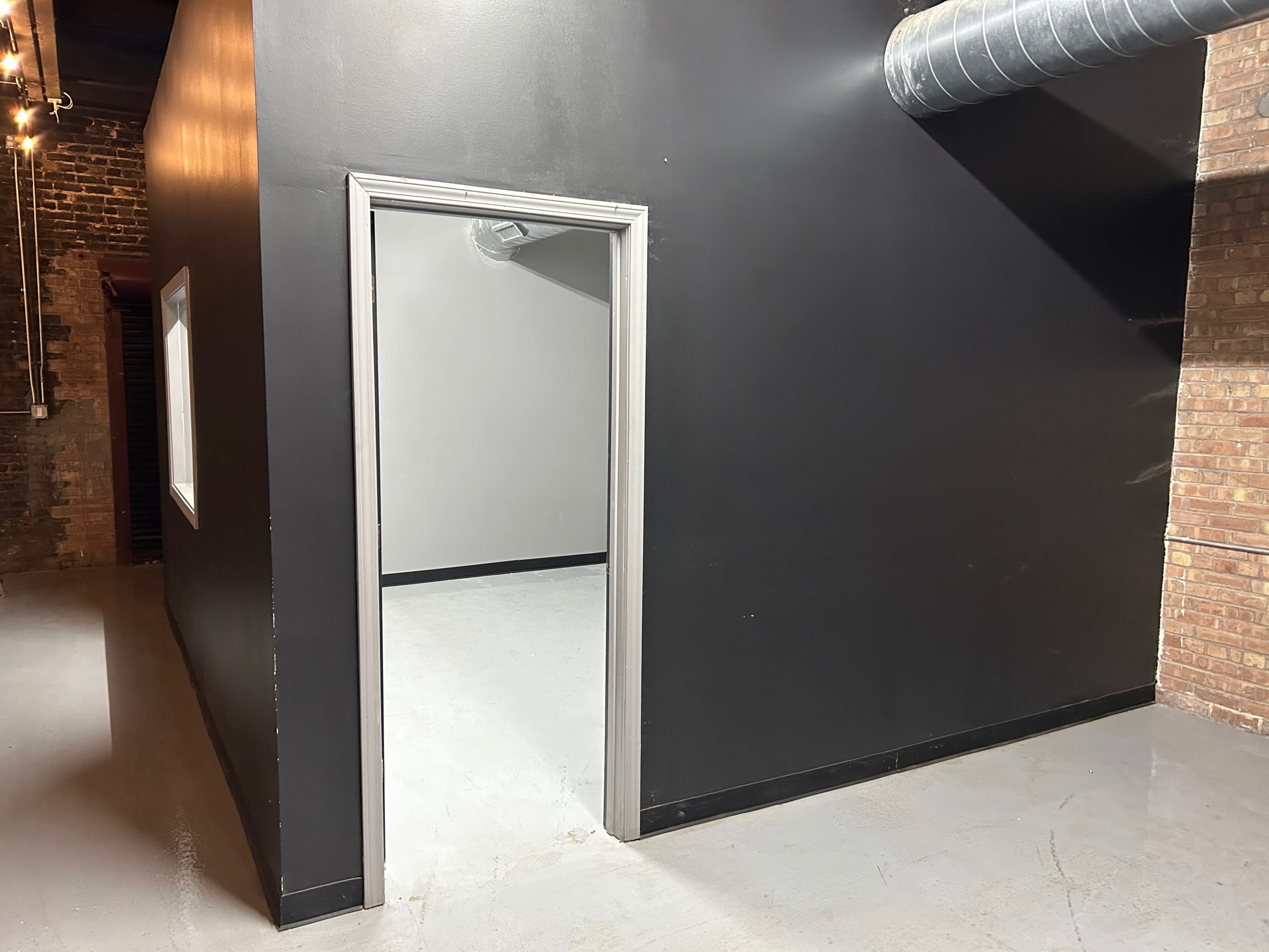 An empty room with a black wall, a doorframe leading to a lighter space, and exposed ventilation ducts along the ceiling.