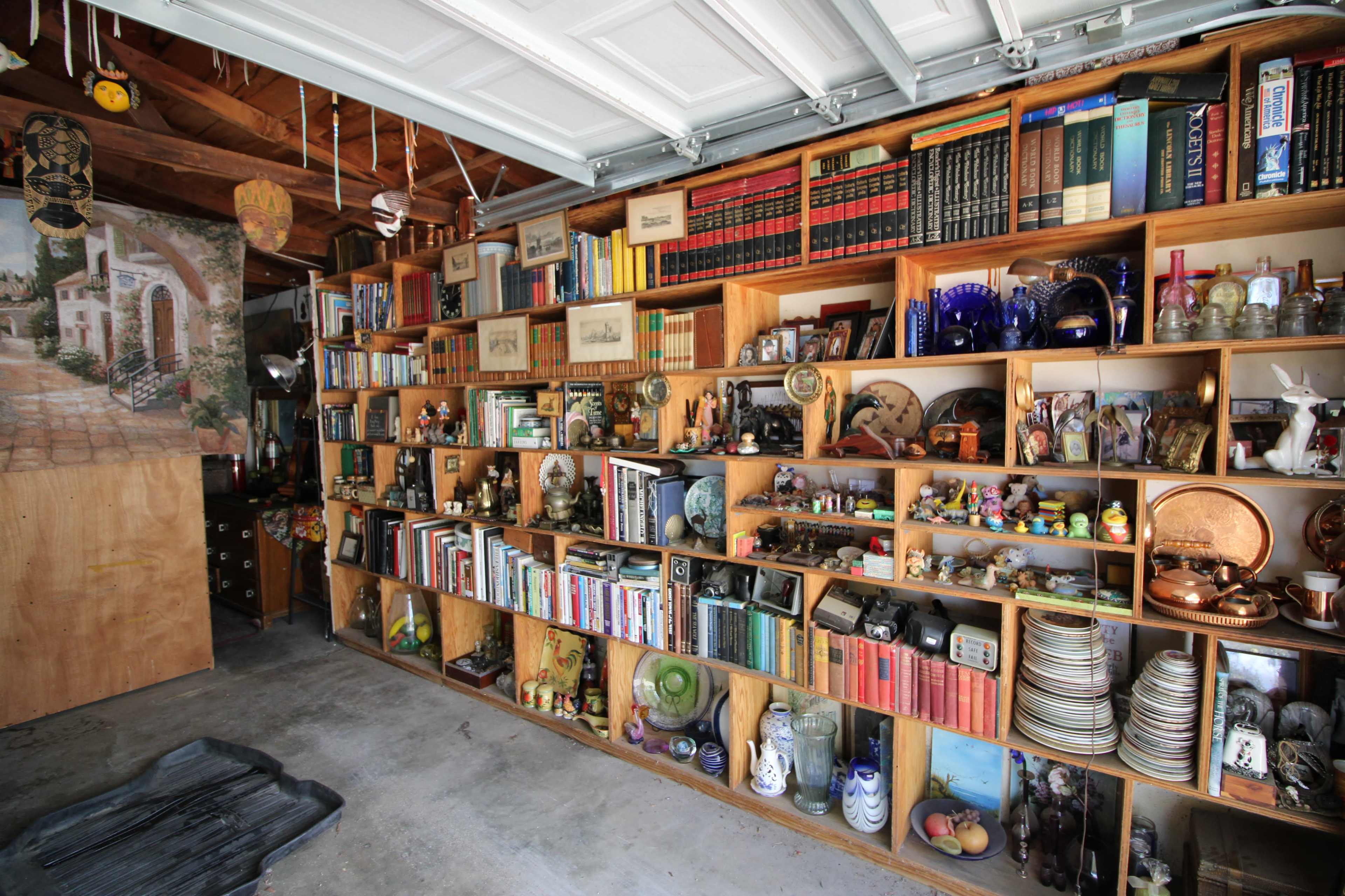 The image shows a garage filled with a wooden shelf stacked with various books, collectibles, and decorative items.