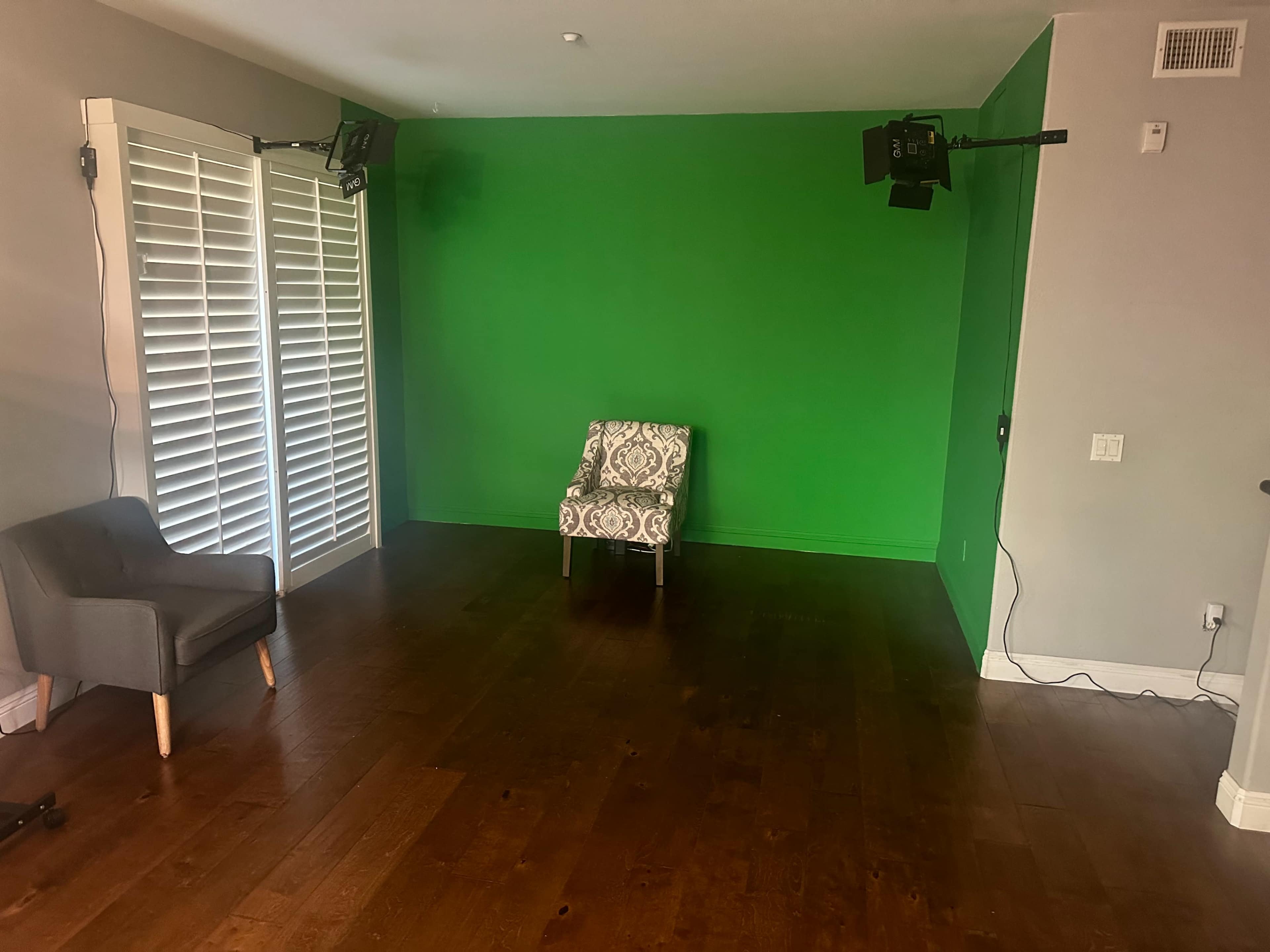 A room with a green-painted wall, a patterned chair in the center, and a gray chair in the corner, with lights mounted on the ceiling.
