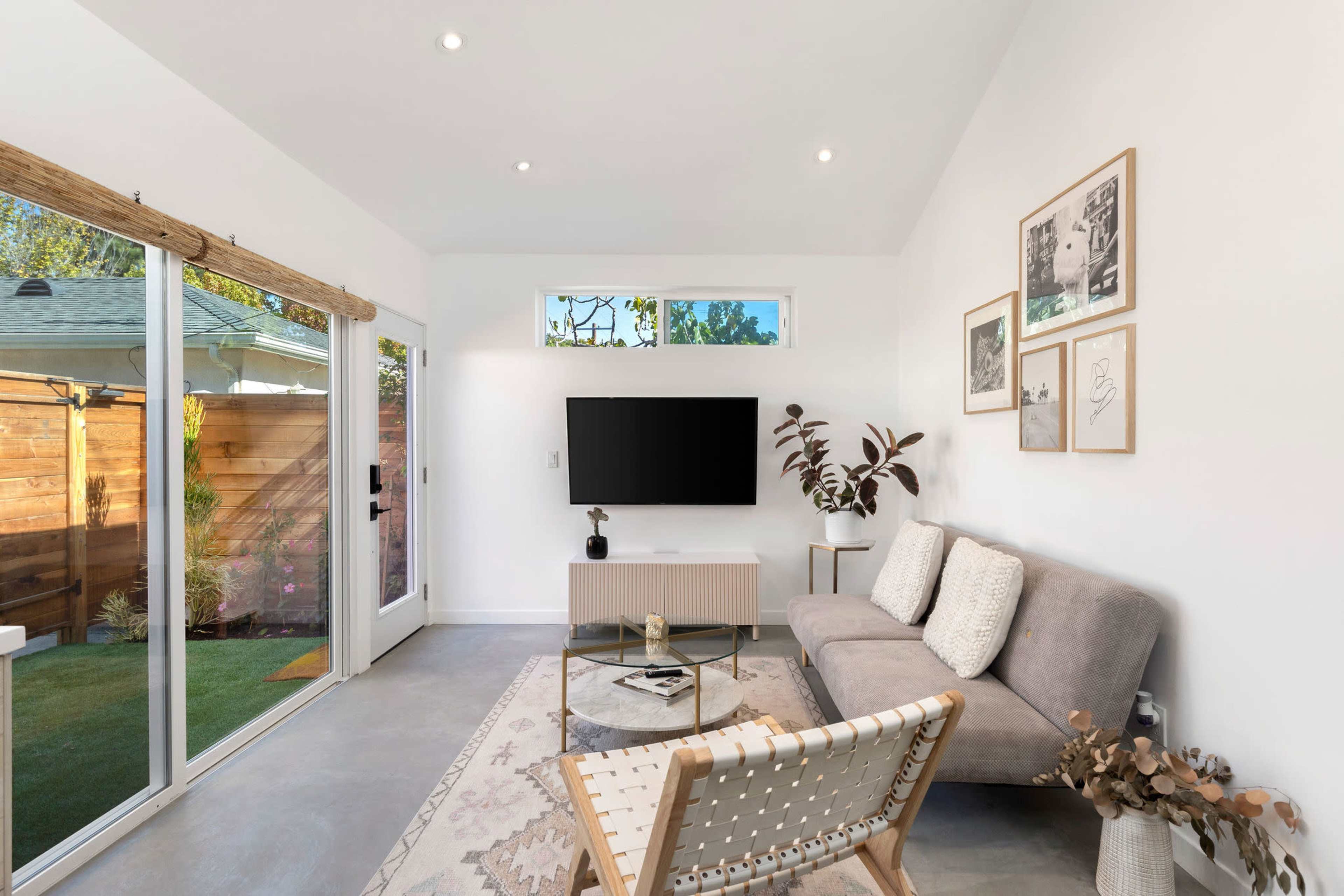 The scene shows a bright living room featuring a gray sofa, a wooden coffee table, a wall-mounted television, and large glass doors leading to an outdoor space.