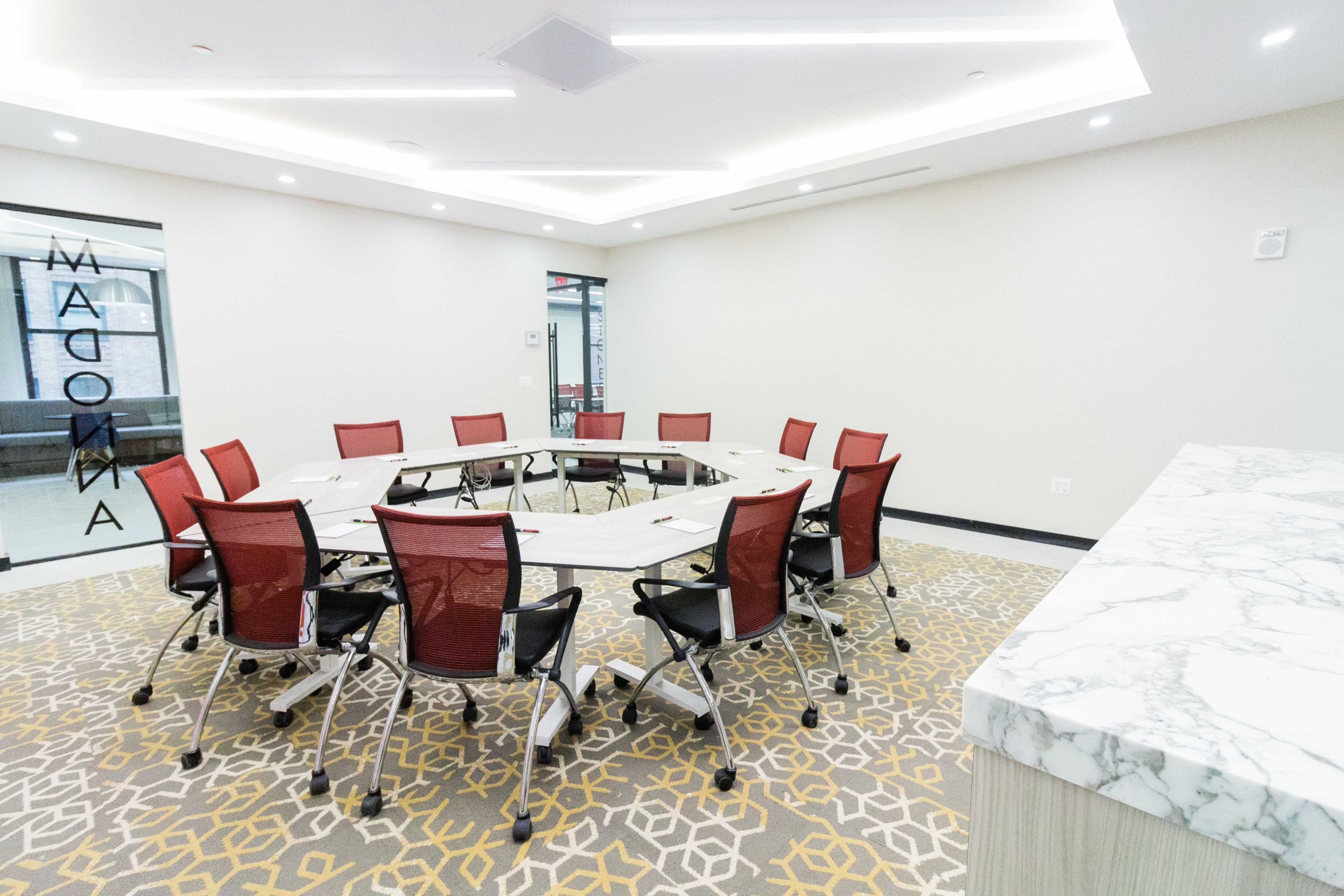 A spacious conference room features an octagonal table surrounded by red mesh chairs, with a patterned carpet and a marble surface nearby.