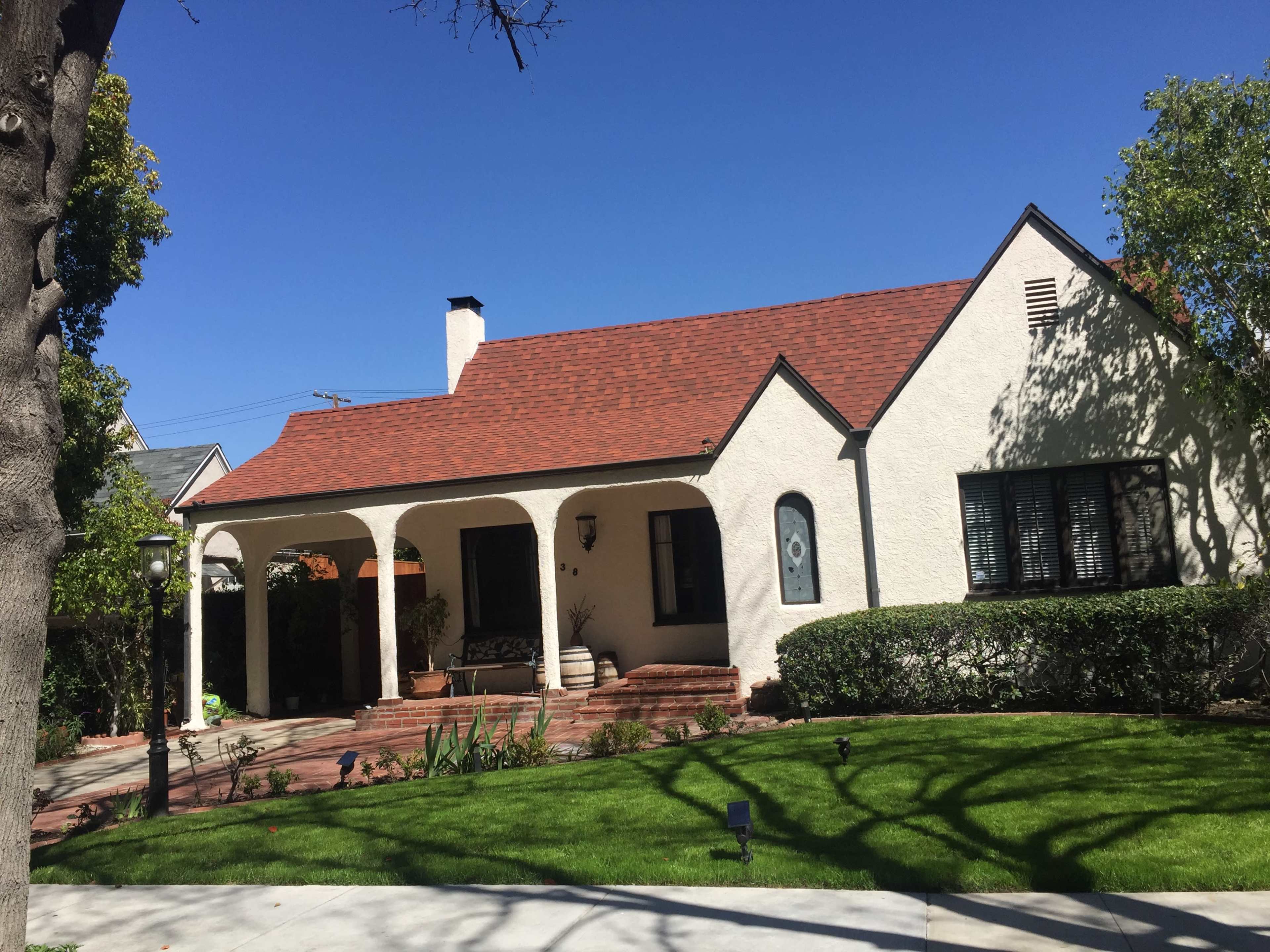 A single-story, white stucco house with a red tile roof features a covered porch supported by arched columns and a manicured lawn.