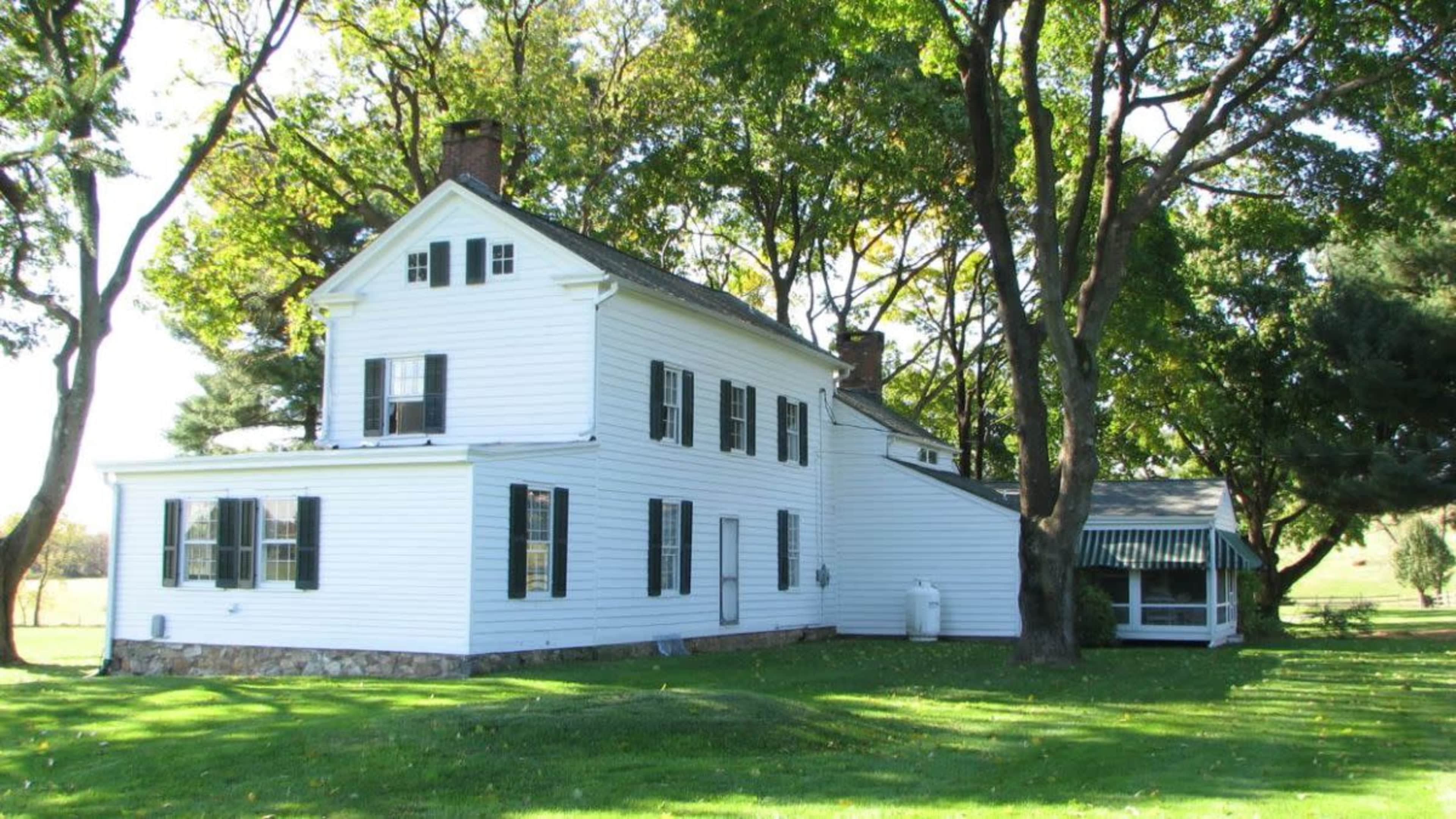 A two-story white house with black shutters is situated under several large trees in a grassy area.