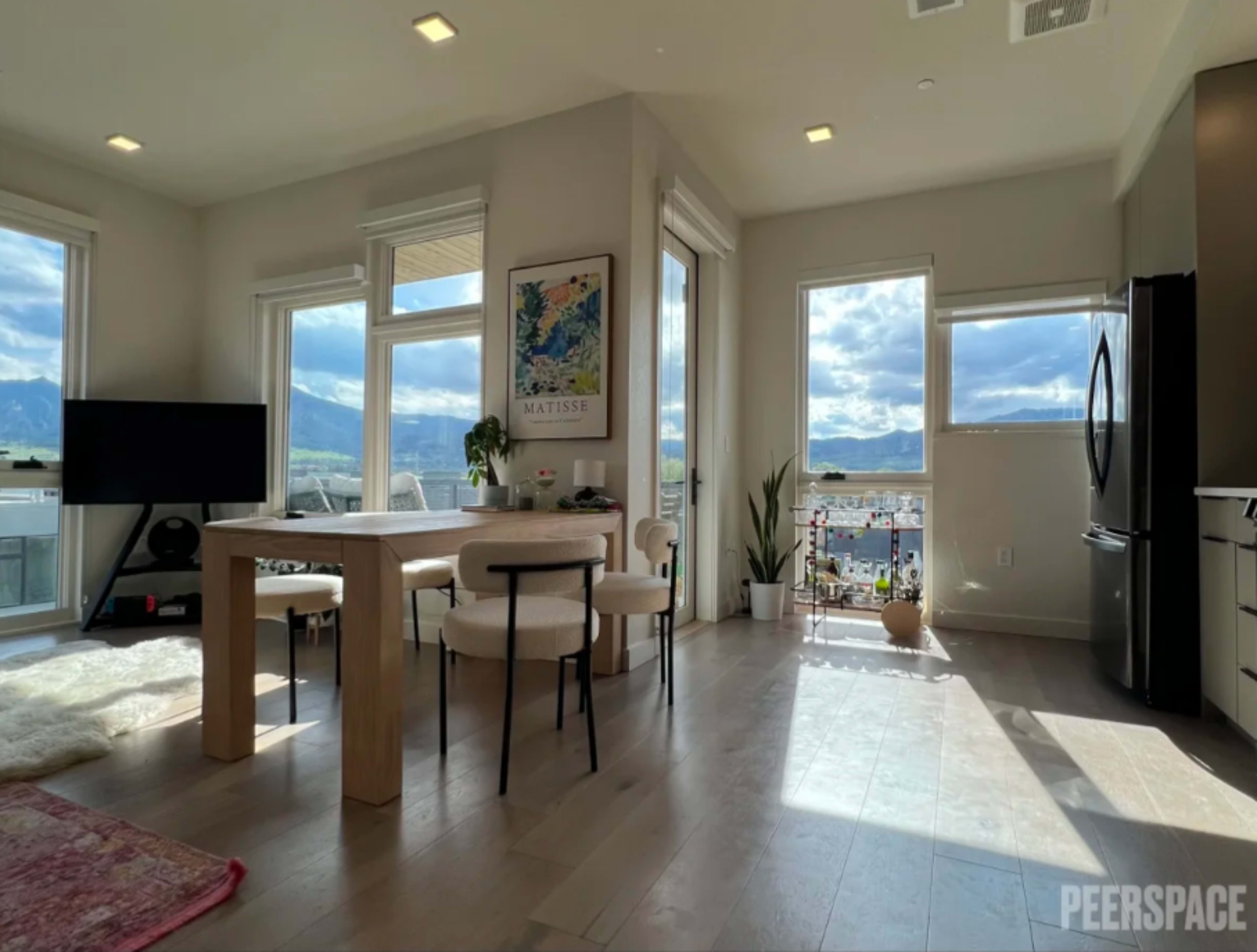 A bright, modern living space with large windows, a dining table with two chairs, a television, and a well-decorated kitchen area.