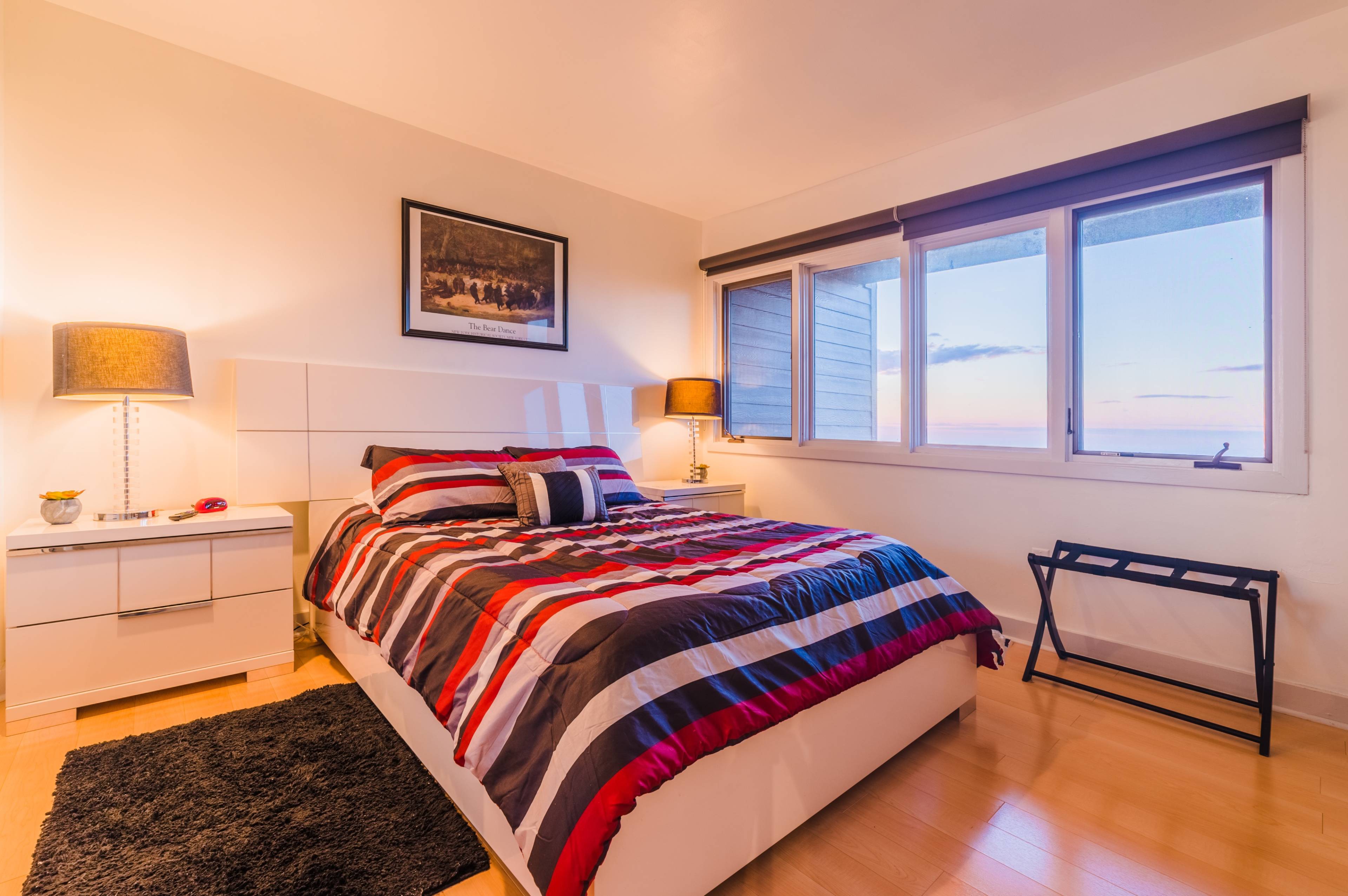 A bedroom featuring a striped bedspread, a nightstand with a lamp, and large windows overlooking a sunset.