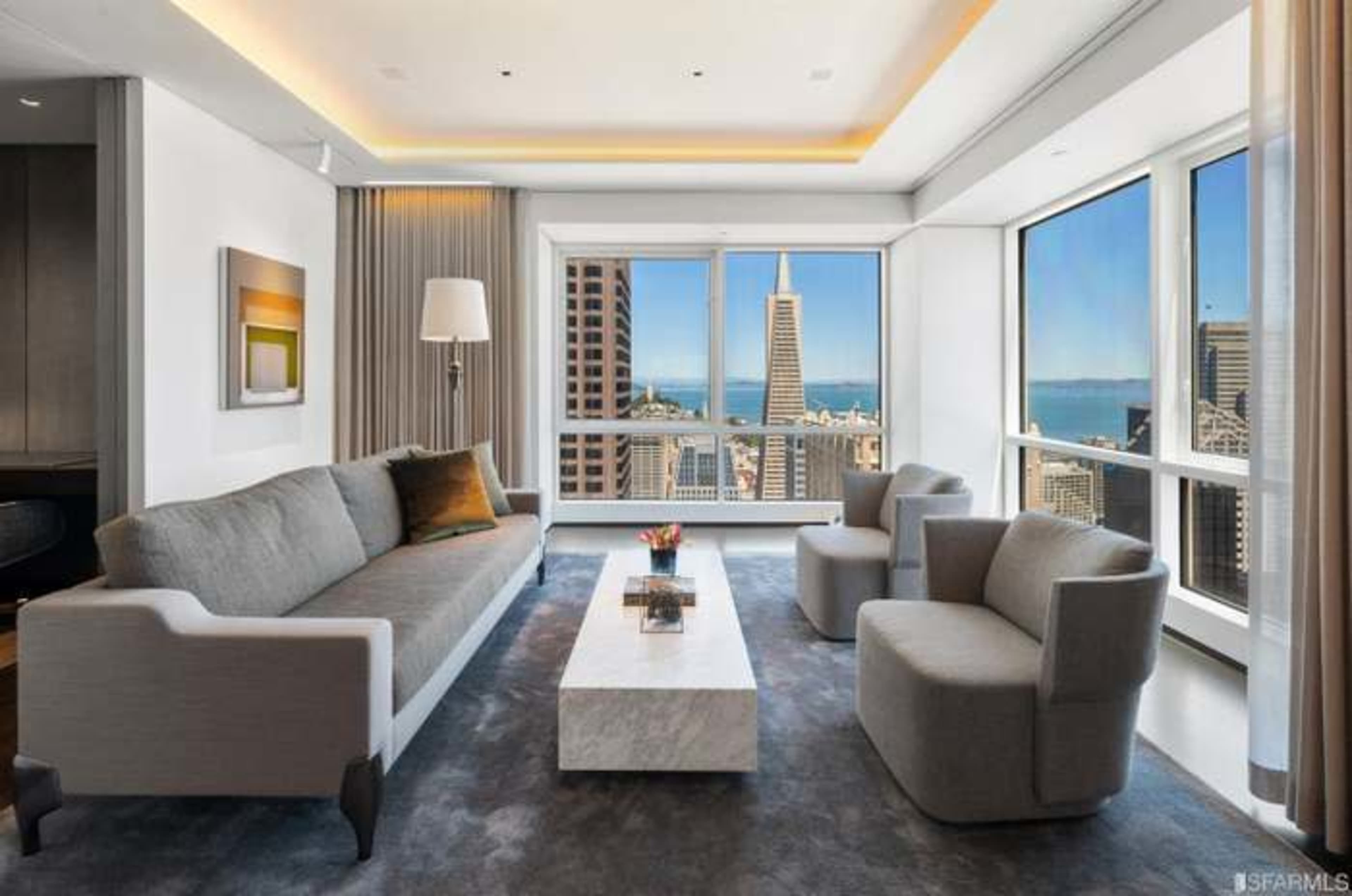 A modern living room features a gray sofa and two armchairs, with a marble coffee table and large windows offering a view of the San Francisco skyline.