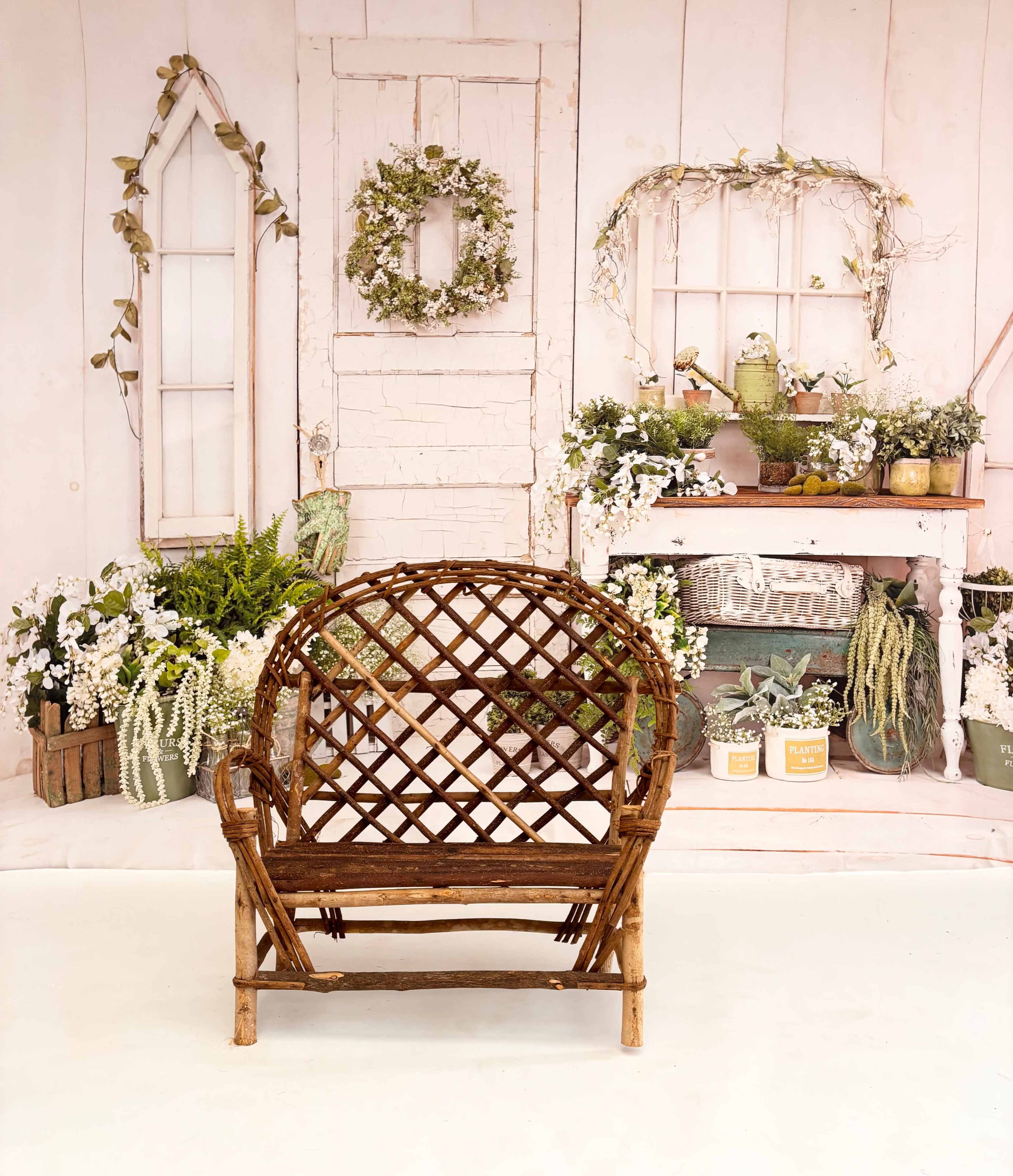 The image features a rustic wicker chair facing a backdrop of a white wooden door and decorated shelves with various plants and floral arrangements.