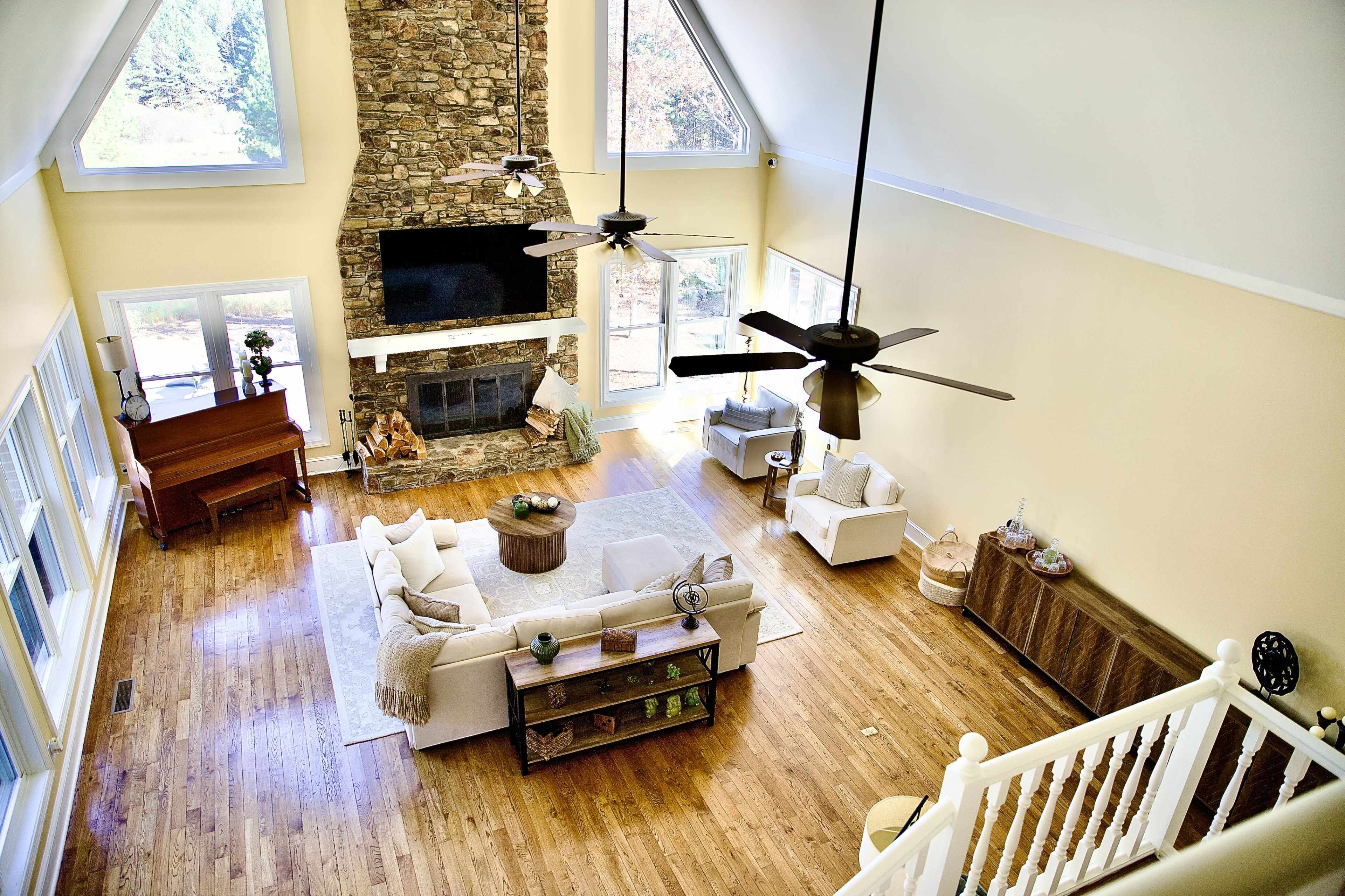 The image shows a spacious living room featuring large windows, a stone fireplace, a flat-screen TV, and a mix of furniture including a piano, sofas, and a coffee table.
