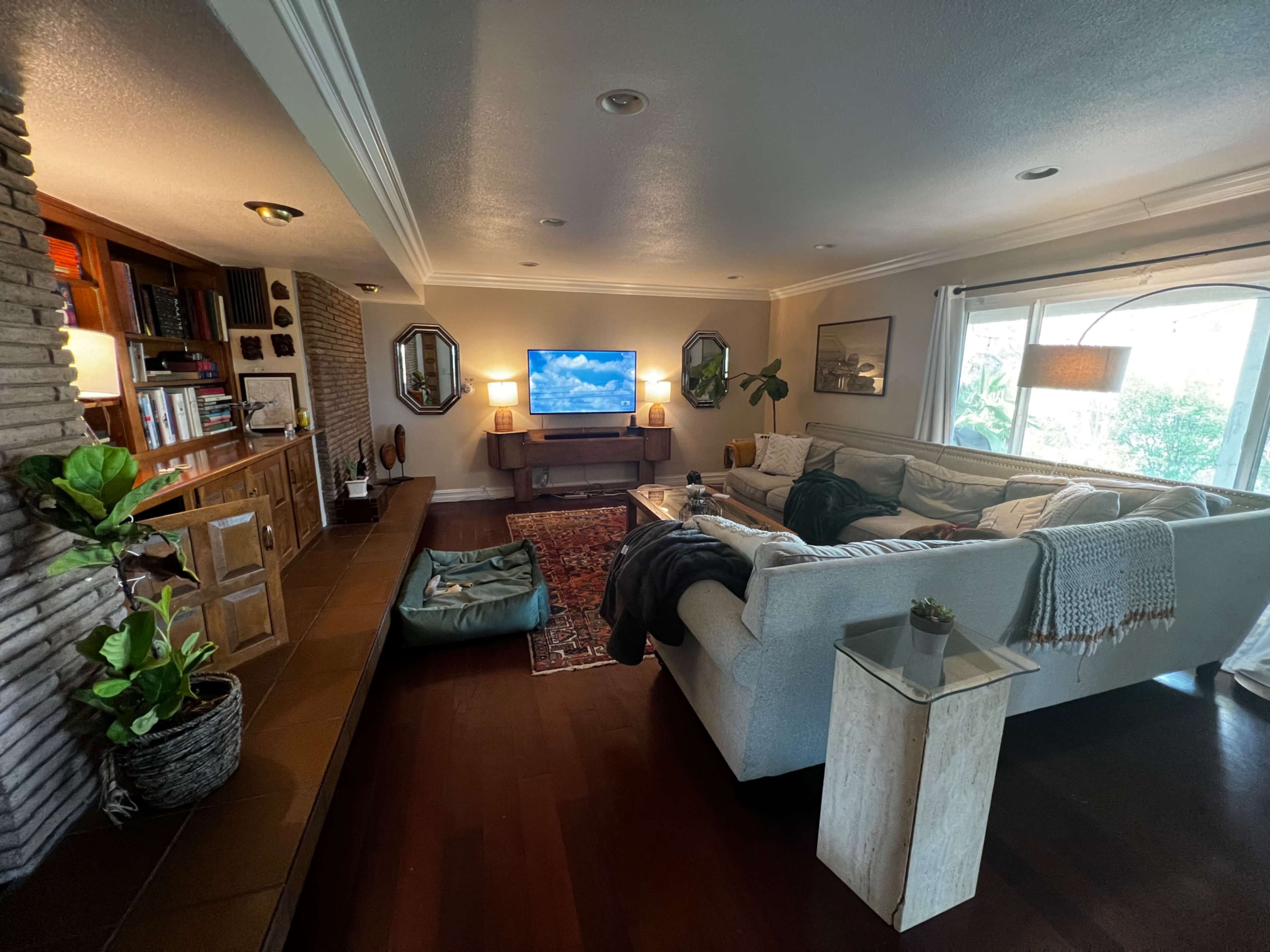 A spacious living room with a large sofa, a coffee table, and a television mounted on the wall, surrounded by plants and decorative elements.