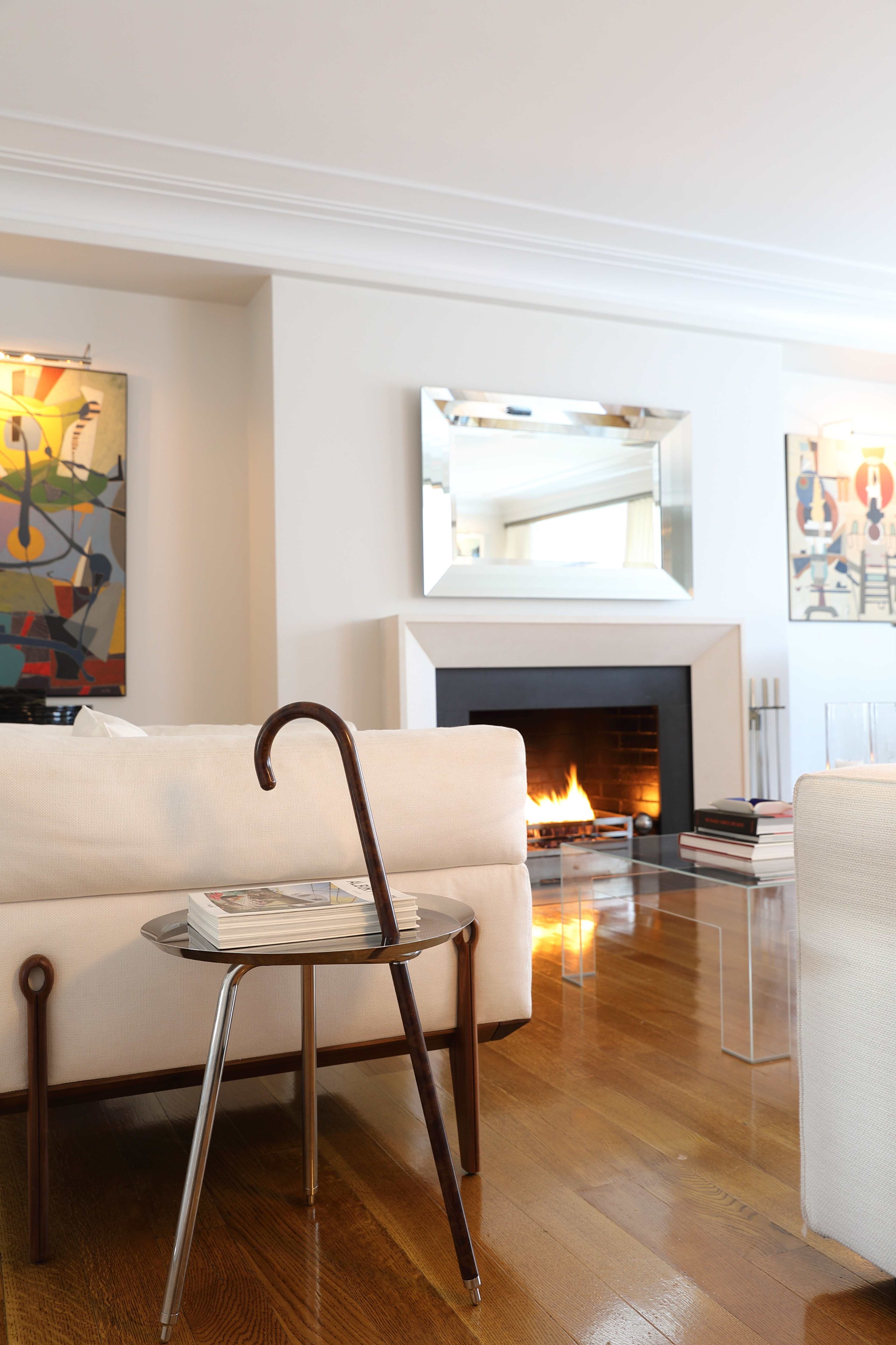 A modern living room features a fireplace, a glass-topped side table with a cane handle, and colorful artwork on the walls.