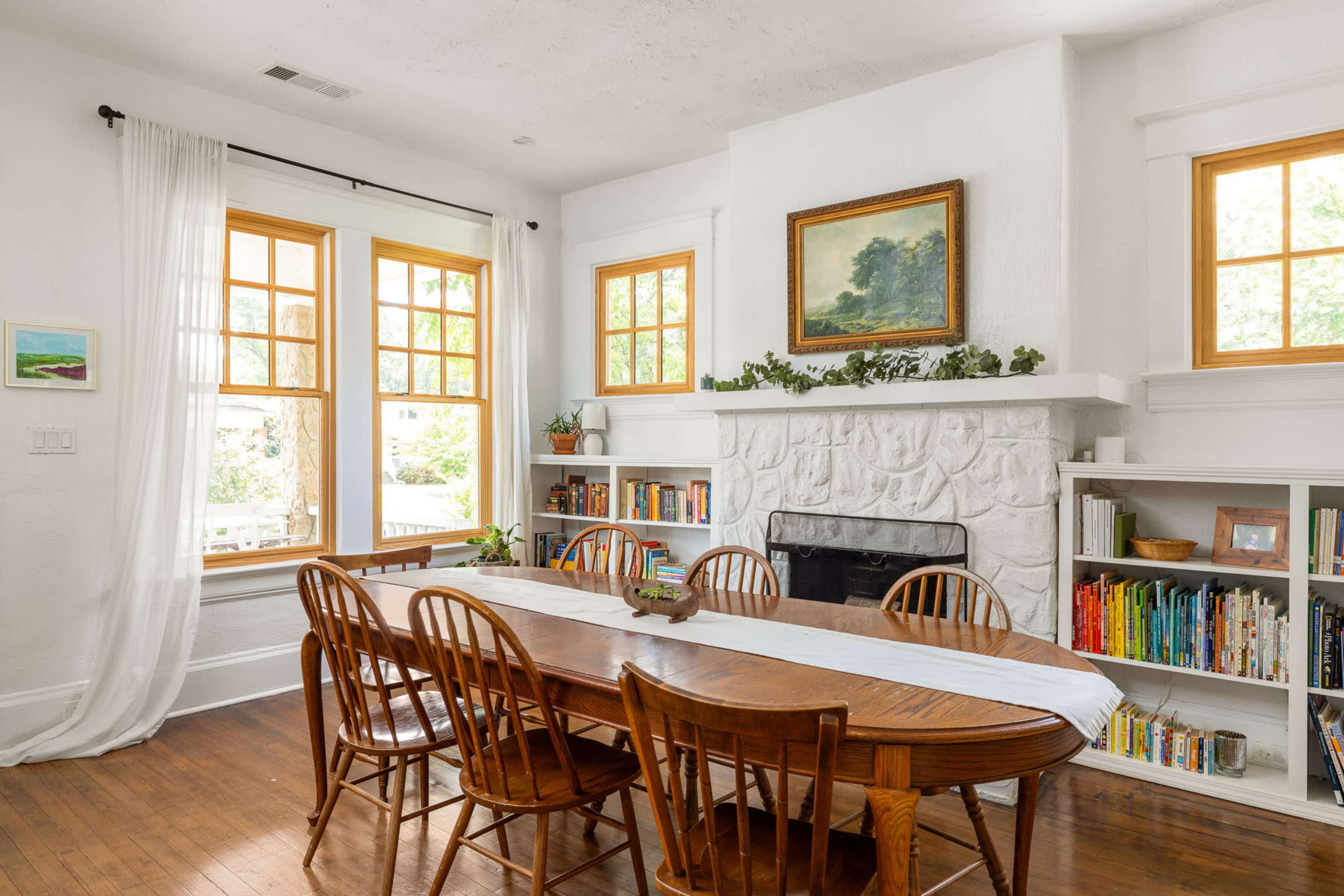 A dining room features a long wooden table surrounded by eight chairs, with large windows and a stone fireplace decorated with a painting and plants.