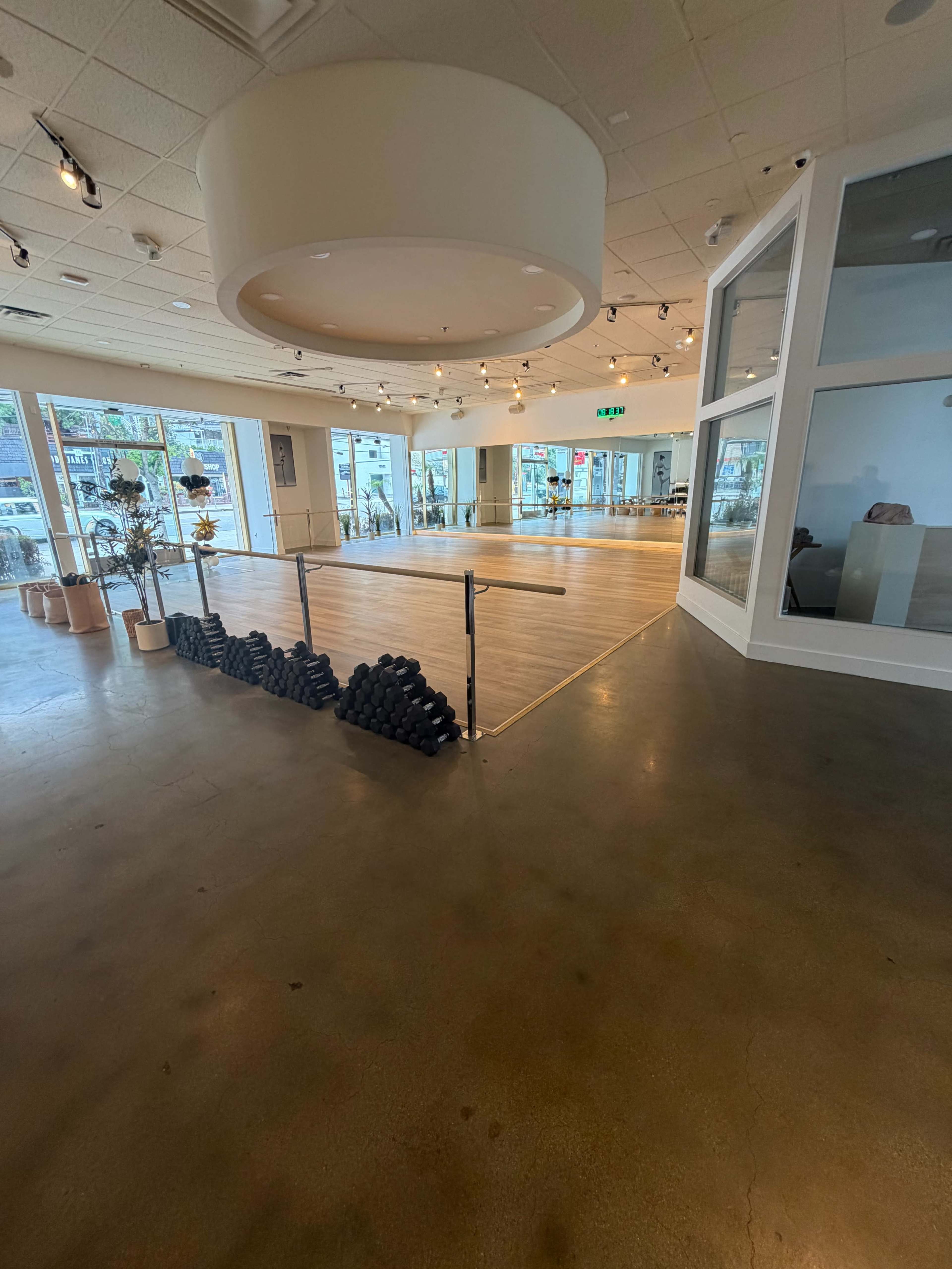 The image shows a spacious, well-lit studio with large windows, a polished wooden floor, and a row of black weights arranged near the entrance.