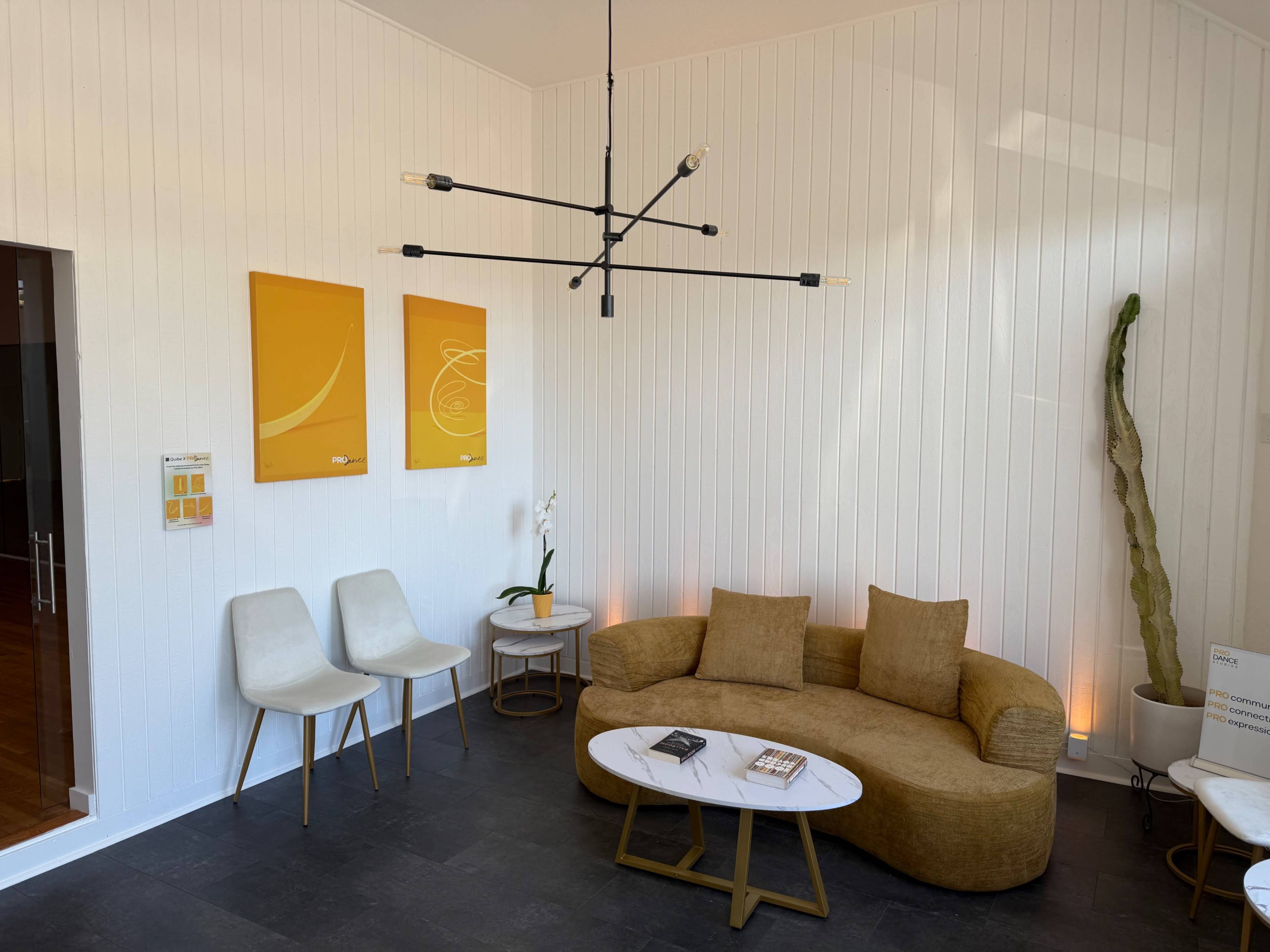 The image shows a modern waiting area with a curved brown couch, two white chairs, a coffee table, and abstract artwork on the walls.