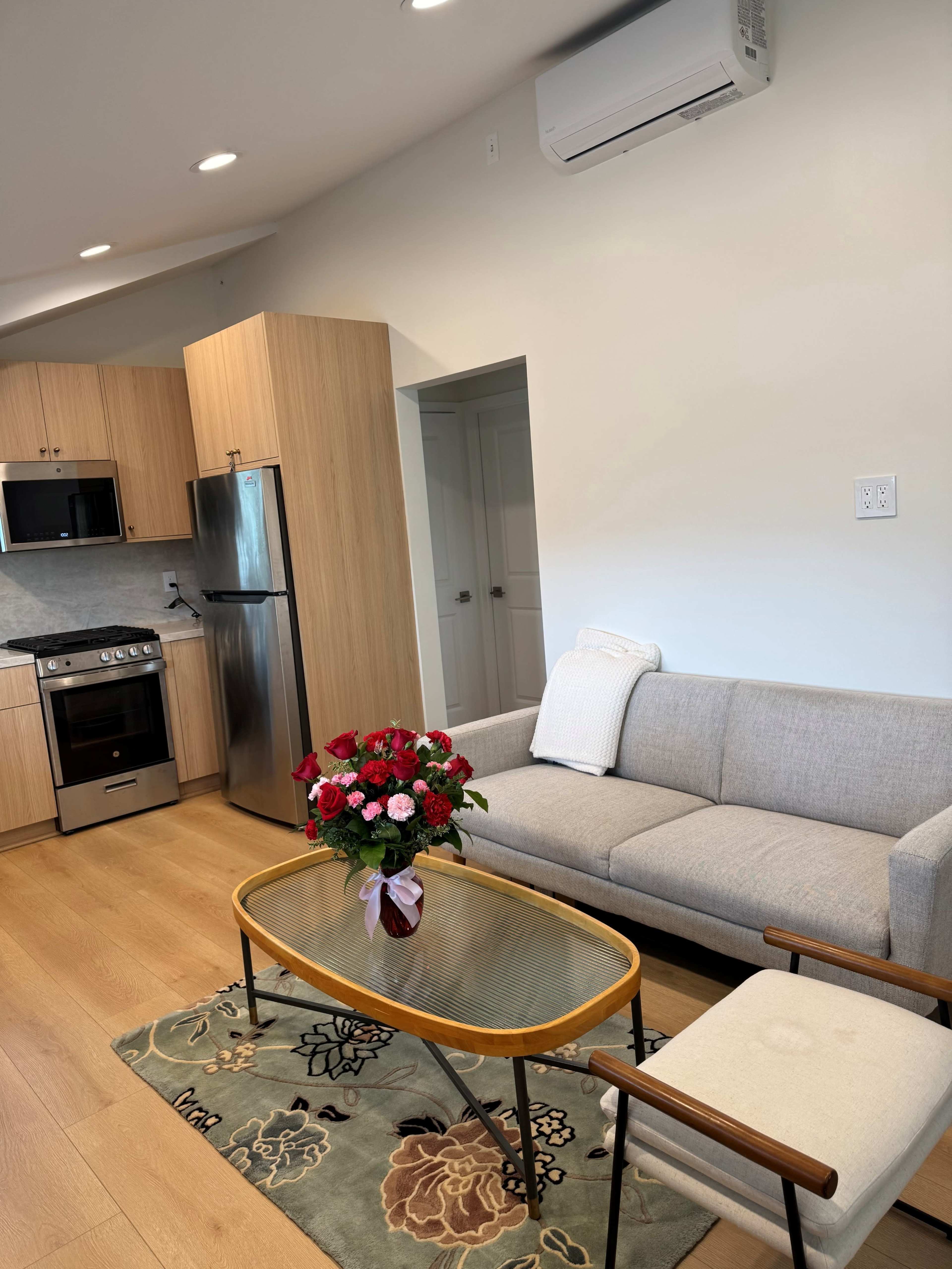 The image shows a modern living area featuring a gray couch, a glass coffee table with a bouquet of red roses, and a kitchen visible in the background with wooden cabinetry and stainless steel appliances.
