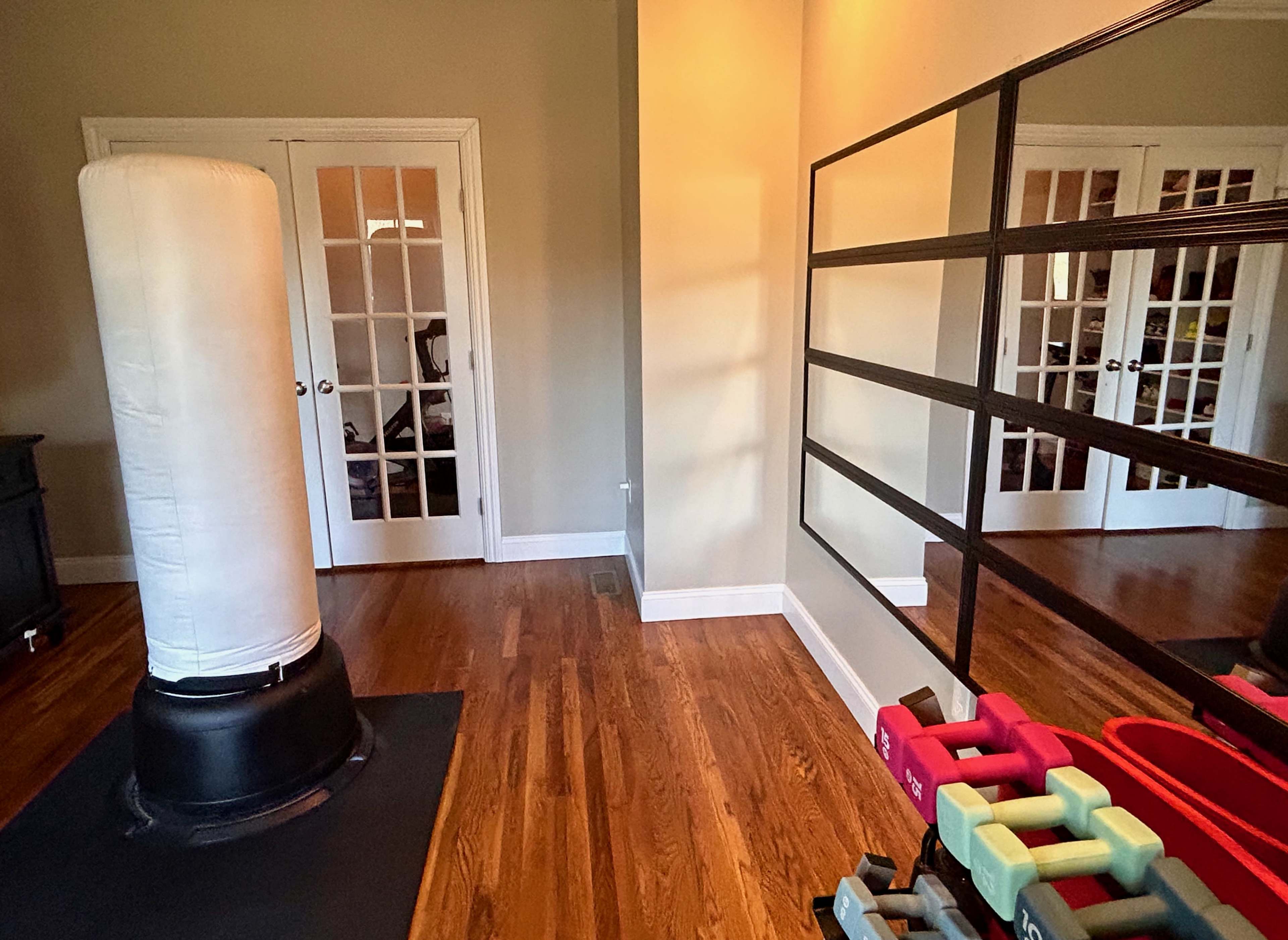 A home gym area featuring a punching bag, a set of dumbbells, and a wall mirror, with double doors leading to another room in the background.