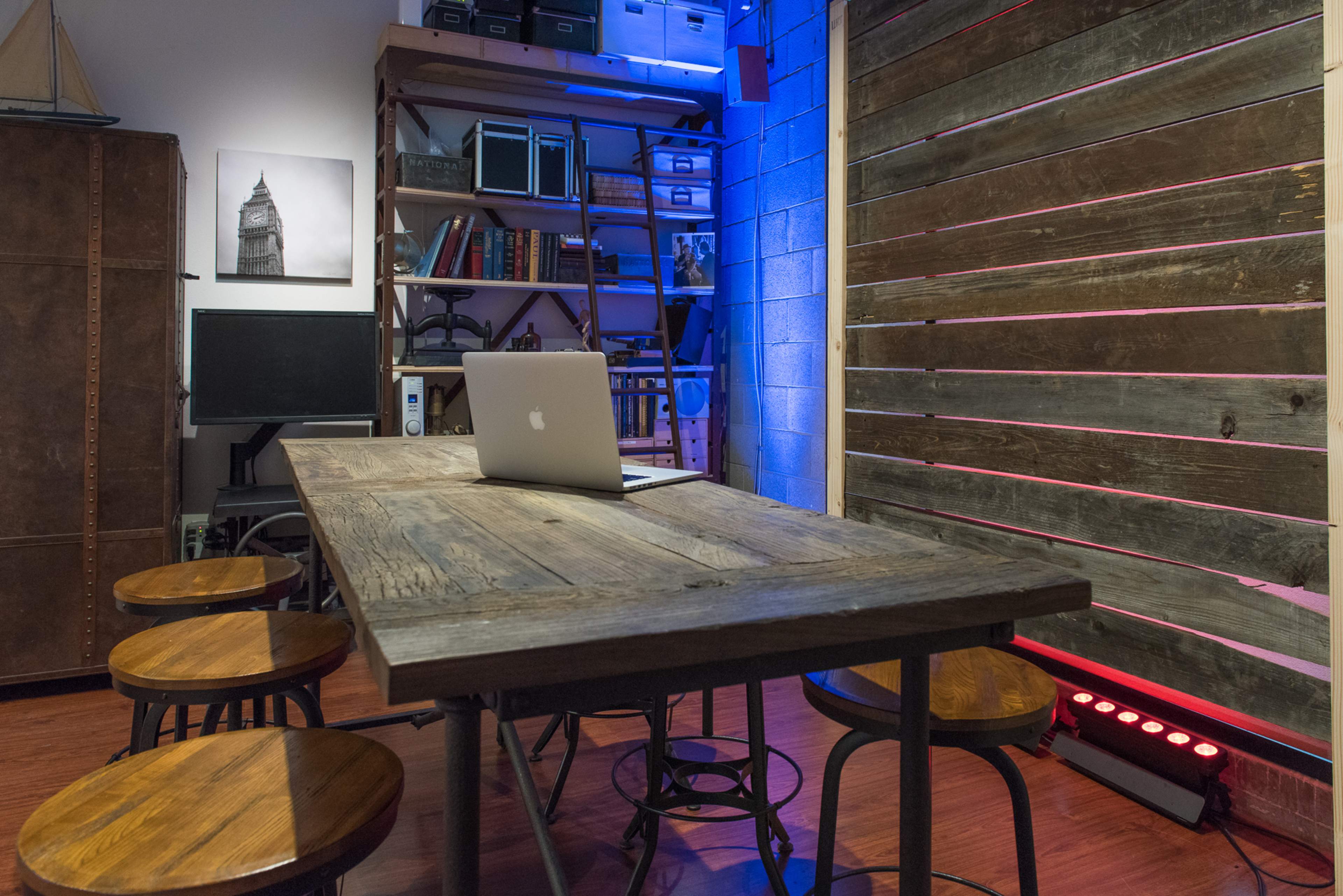 A conference room with a wooden table and four stools, a laptop on the table, and a wall featuring shelves filled with books and decorative items.