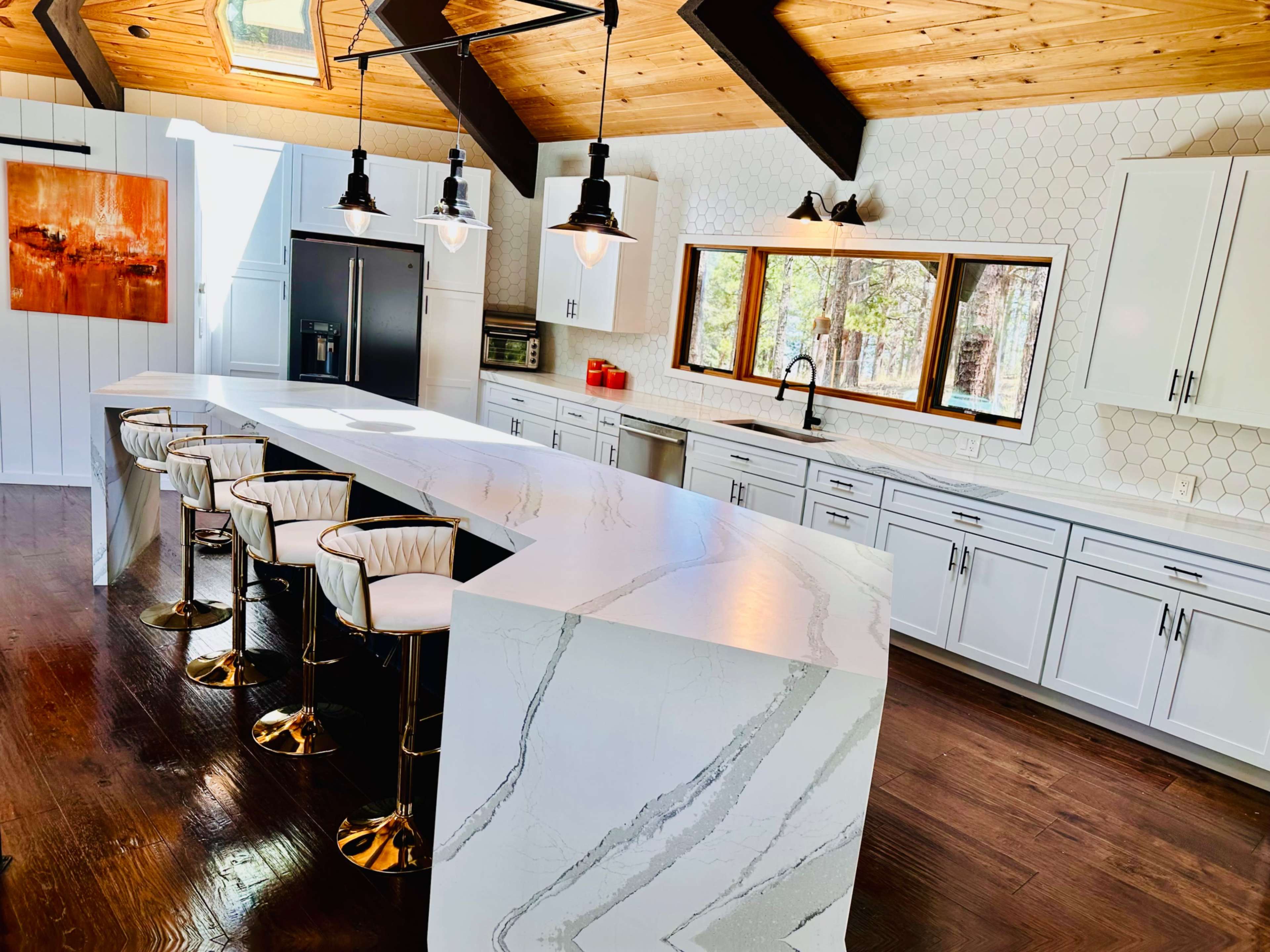 A modern kitchen features a large white countertop with bar stools, a window with natural light, and sleek cabinetry against a wood-paneled ceiling.