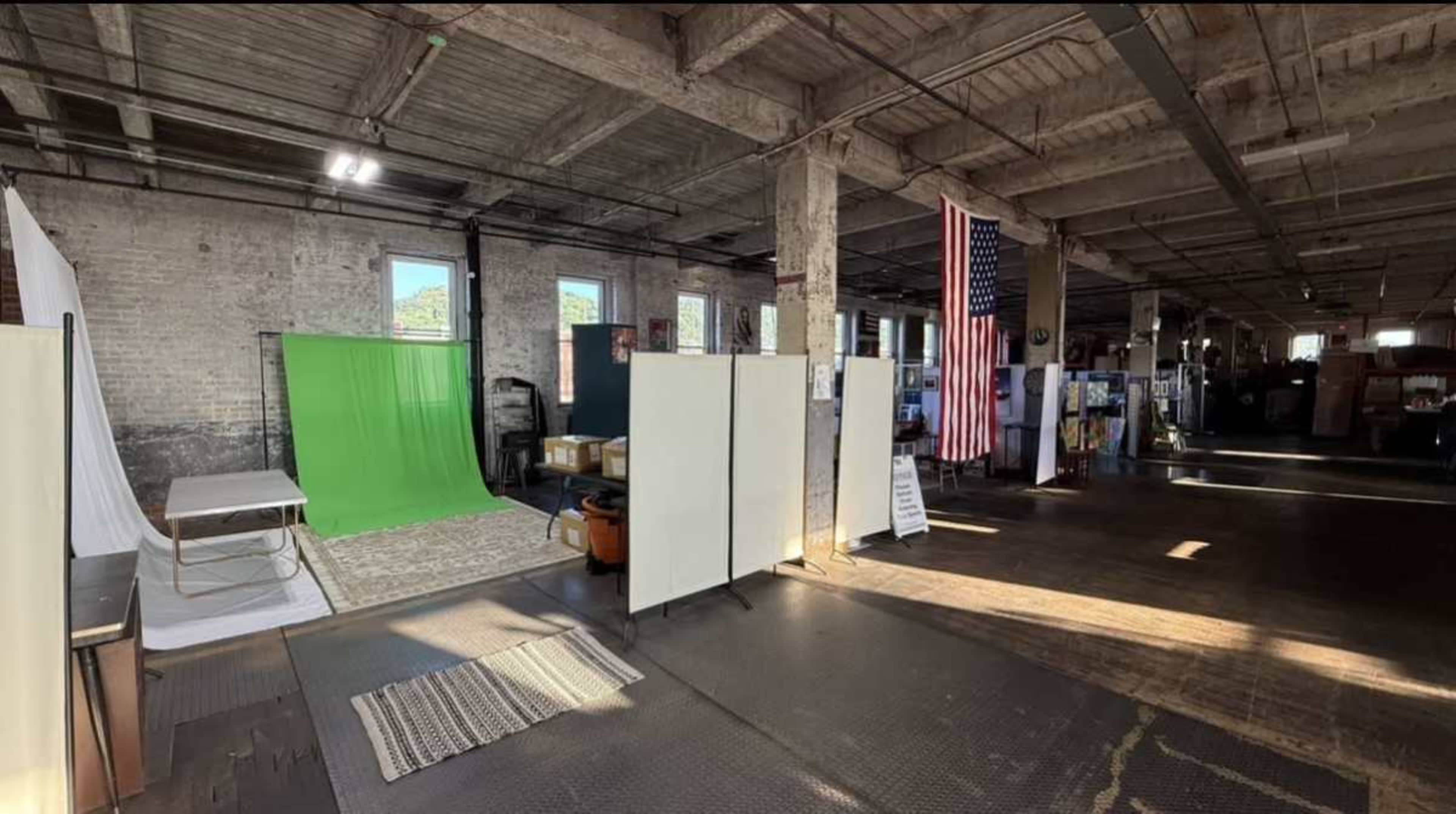 The image shows an industrial space featuring a green screen setup, a white backdrop, an American flag, and various boxes, all illuminated by natural light from large windows.