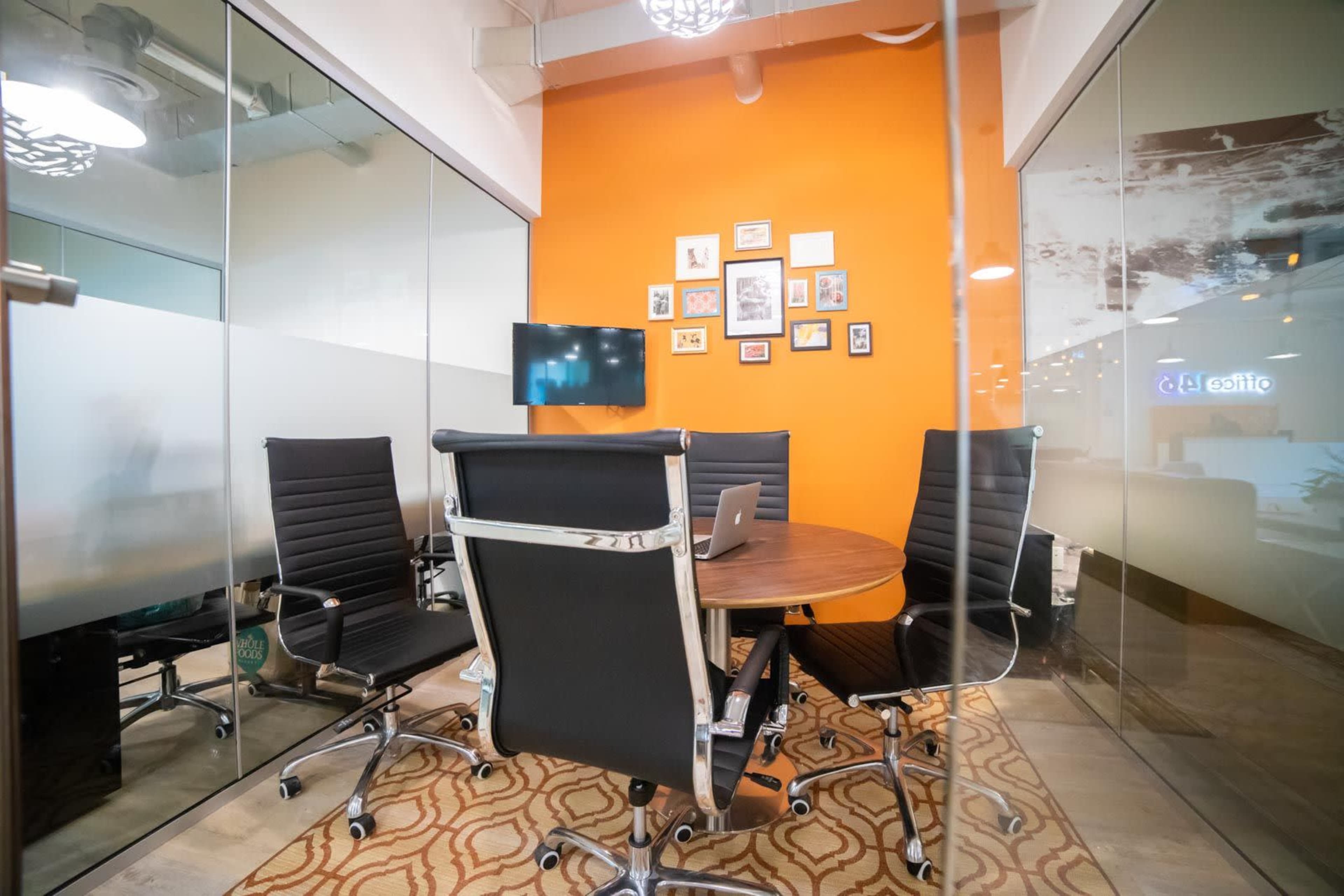 A modern conference room features a round table surrounded by black chairs, with an orange wall displaying framed pictures and a mounted TV.