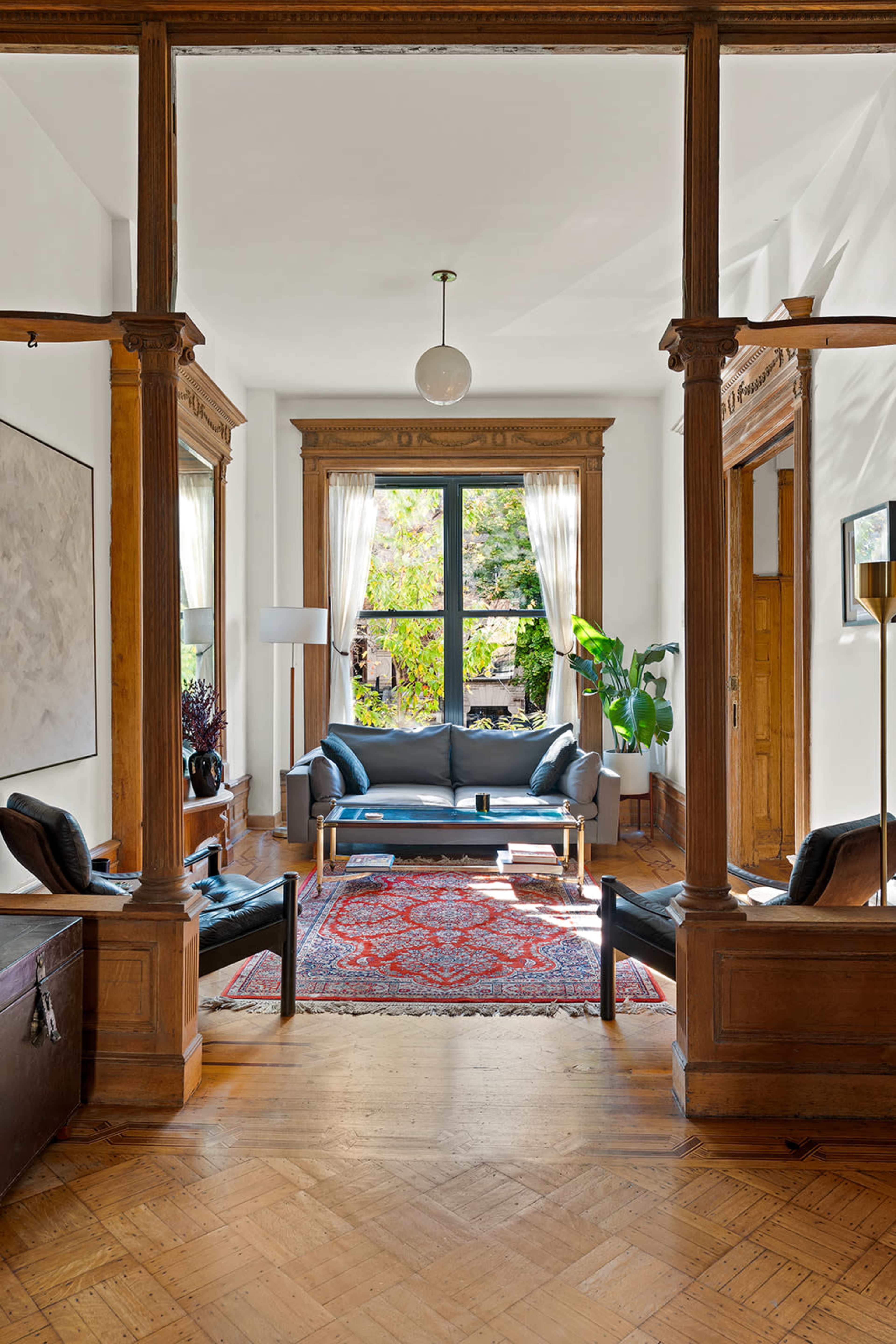The image shows a spacious living room featuring a gray sofa, two chairs, a decorative rug, and large windows with natural light illuminating the space.