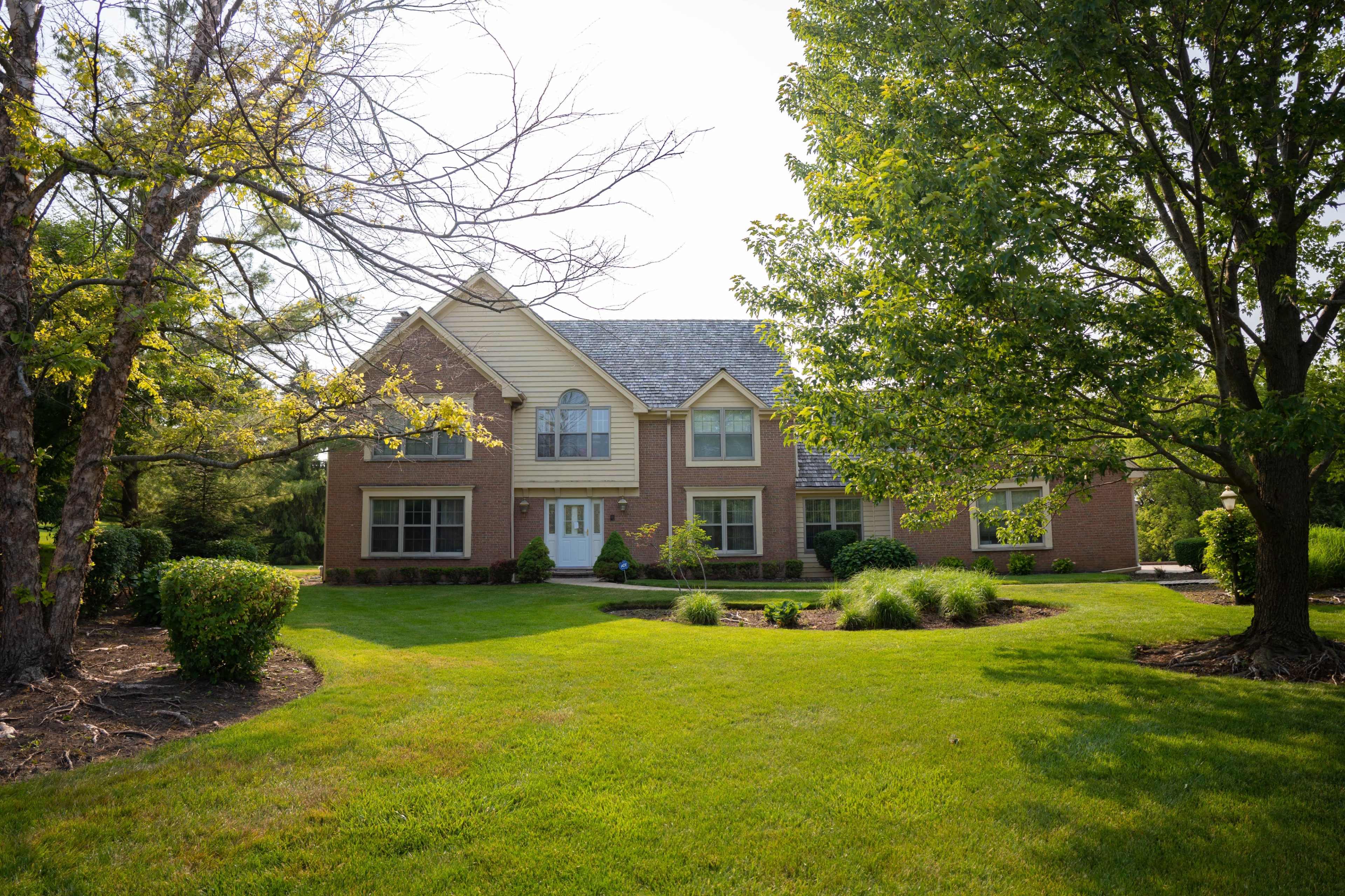 A brick house with a pitched roof is set on a landscaped lawn surrounded by trees and shrubs.