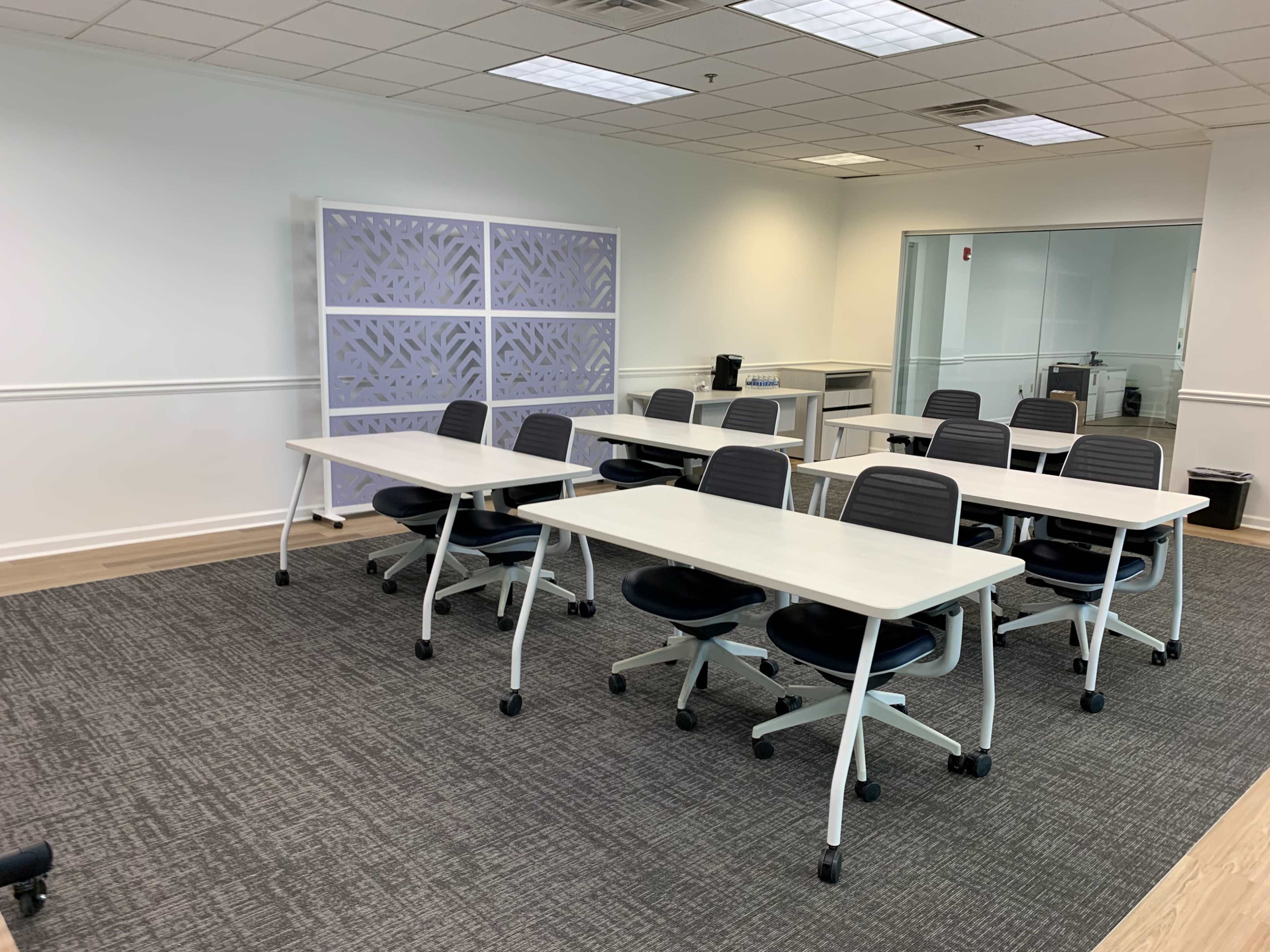 A sparse meeting room features several movable tables and chairs arranged on a carpeted floor.
