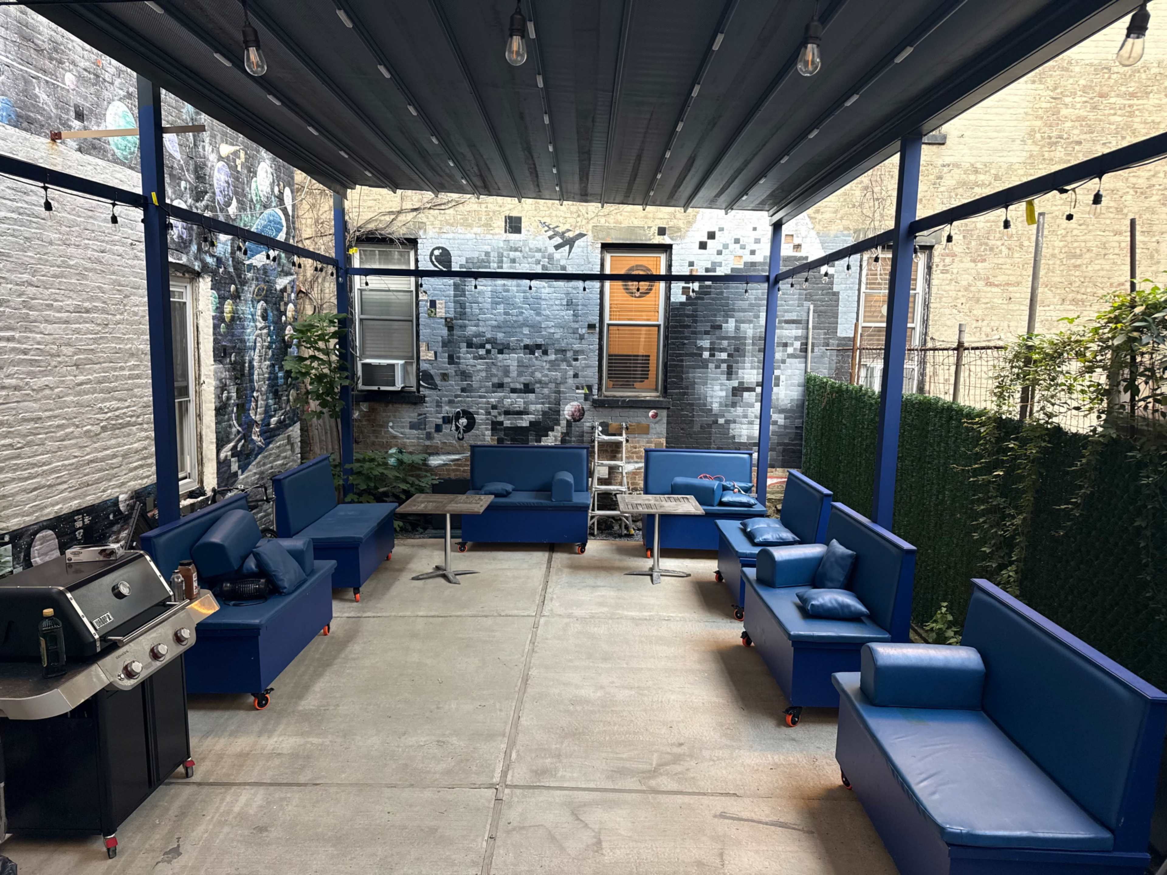 The image shows a cozy outdoor patio area with blue seating, a grill, and a mural on the wall.