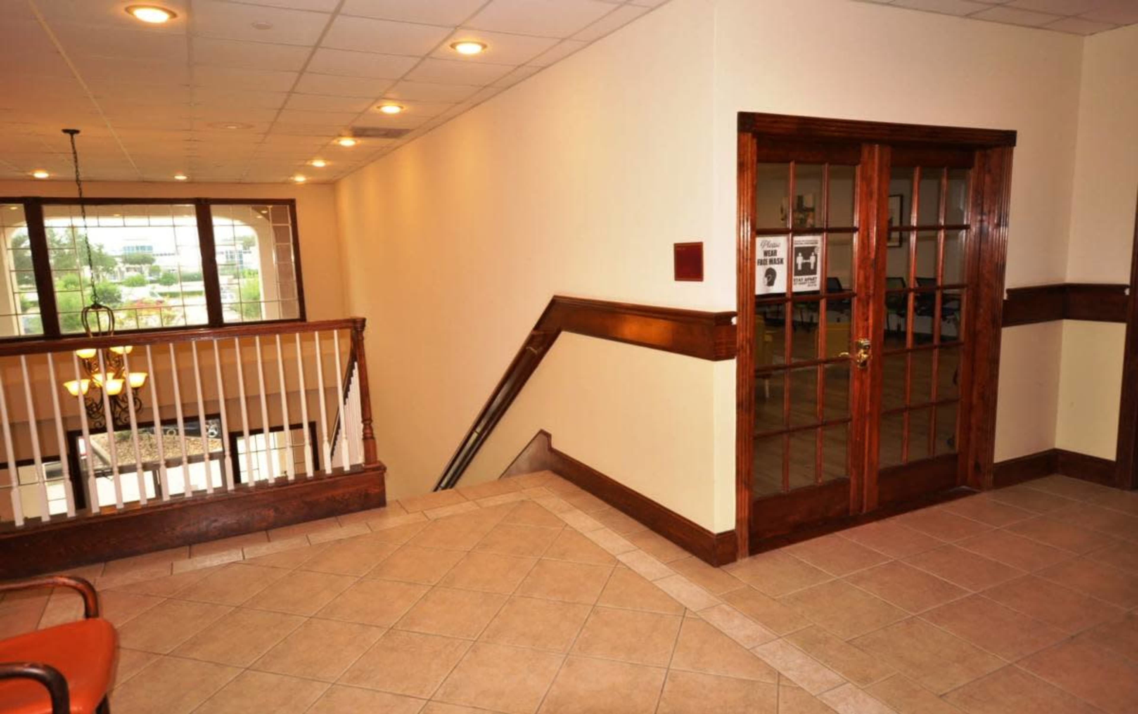 The image shows a well-lit interior hallway with a wooden railing, leading to an open staircase and a pair of glass-paneled doors.
