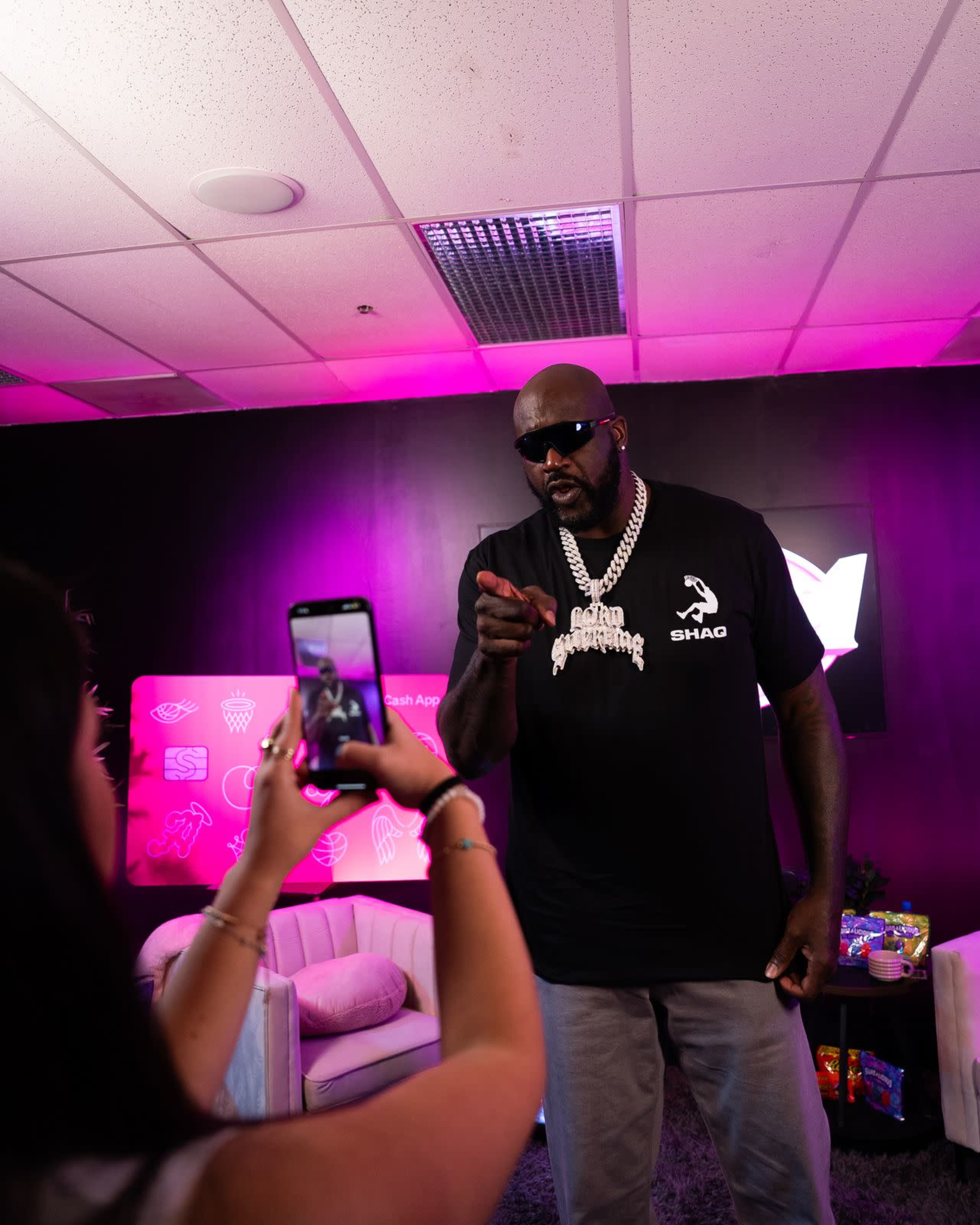 A tall man wearing sunglasses and a black t-shirt is pointing while a person in front of him is taking a photo with a smartphone in a brightly lit room with pink accents.
