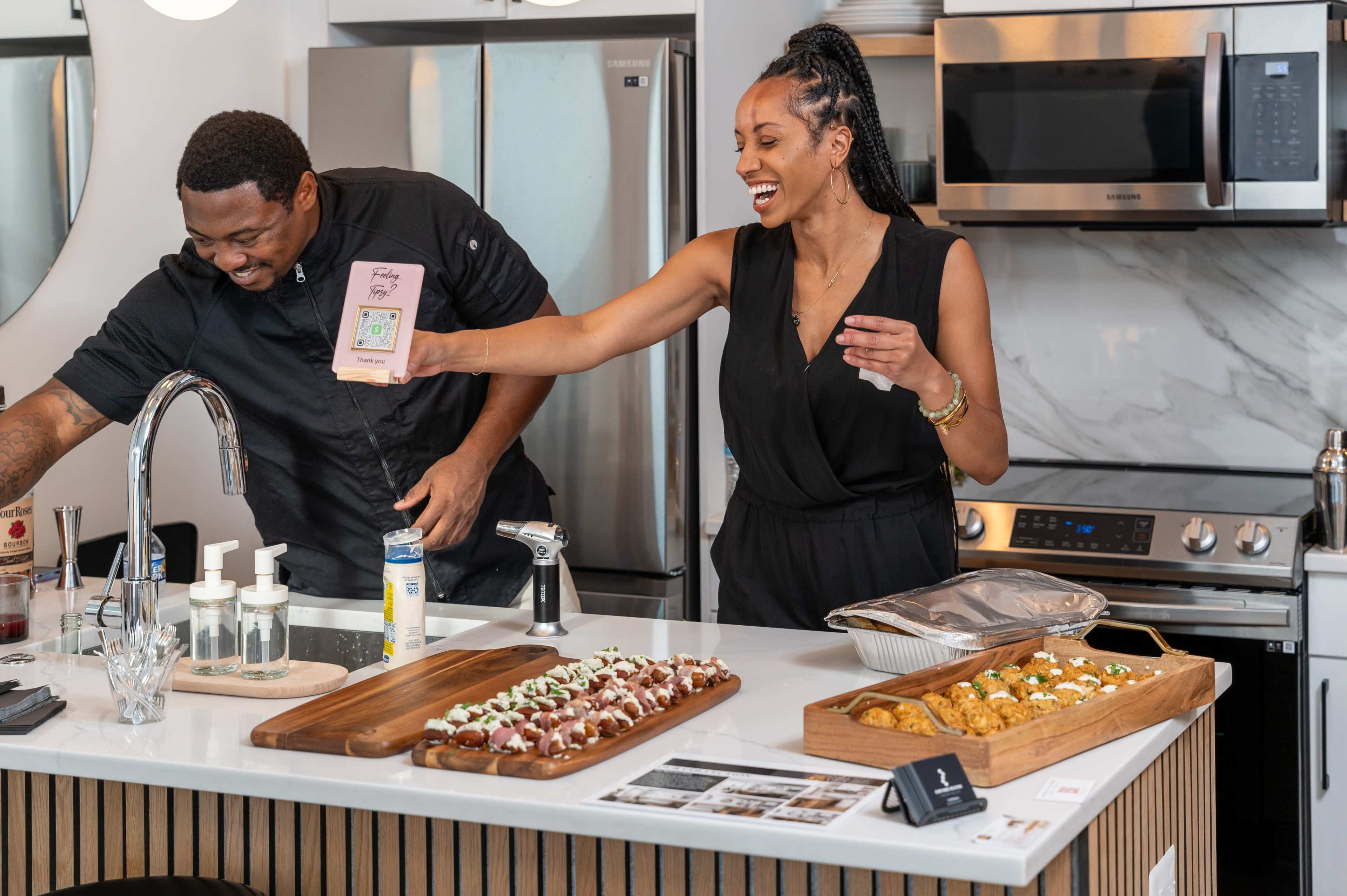 A man and a woman are preparing food in a modern kitchen, with the woman holding a menu and the man working at the sink.