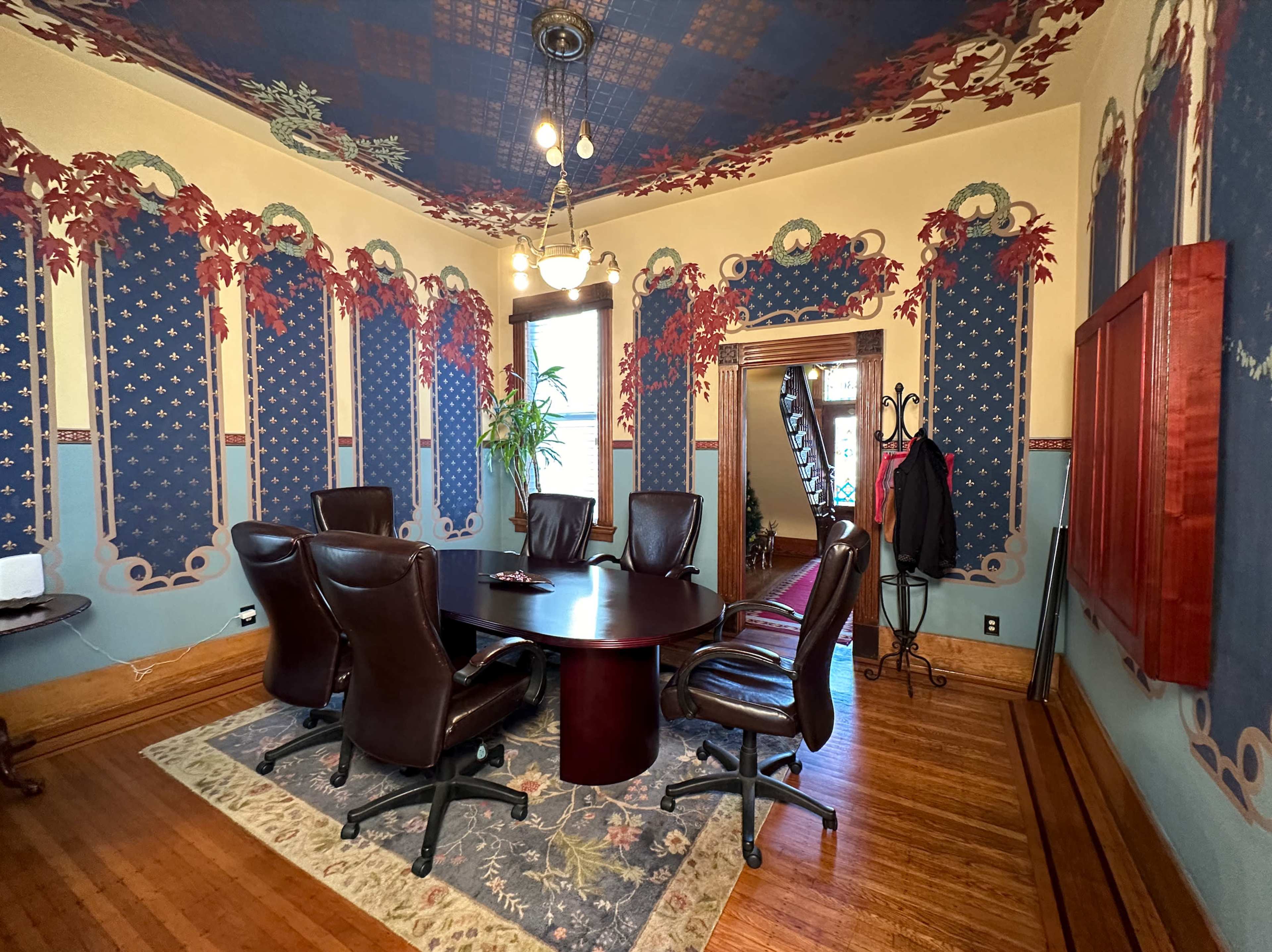 A conference room features a circular table surrounded by six leather chairs, with ornate wallpaper and a patterned rug on the hardwood floor.