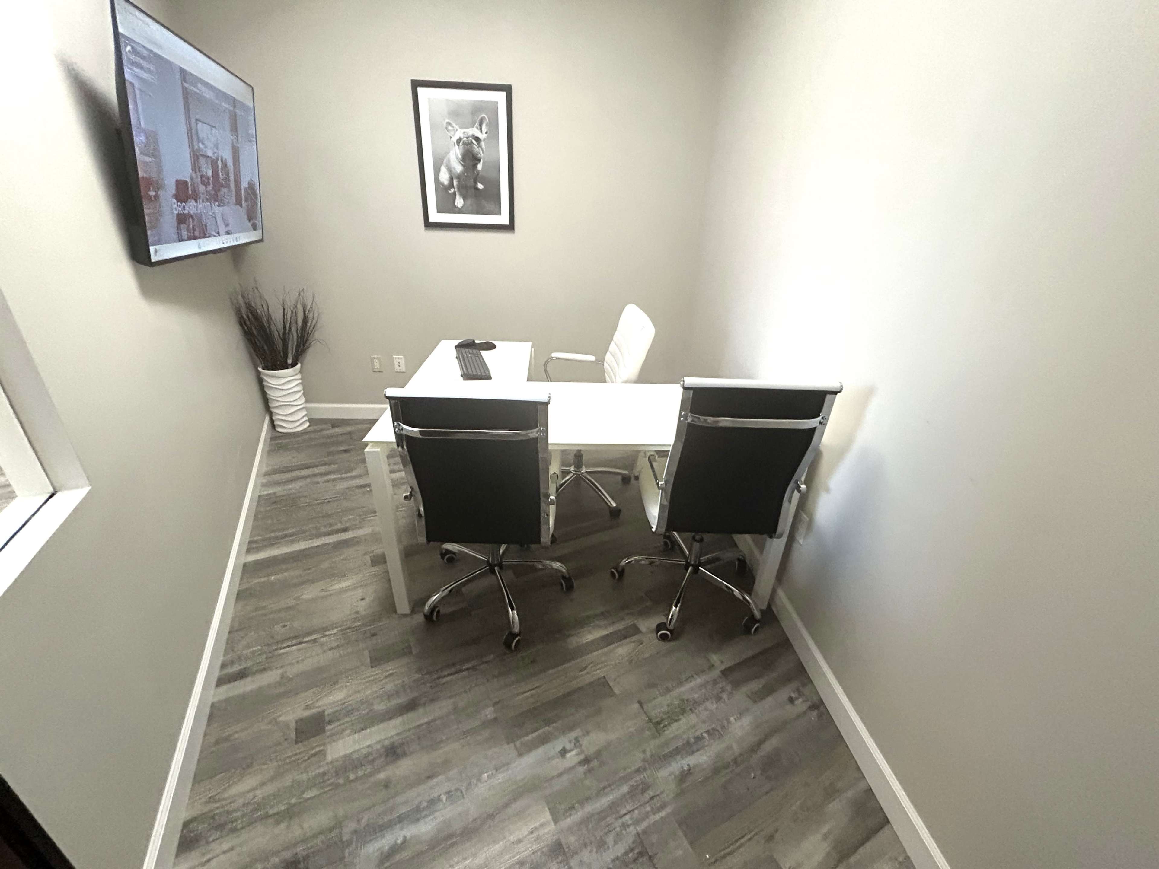 A modern, small office space features a white desk with two chairs, a wall-mounted screen, and a framed picture.