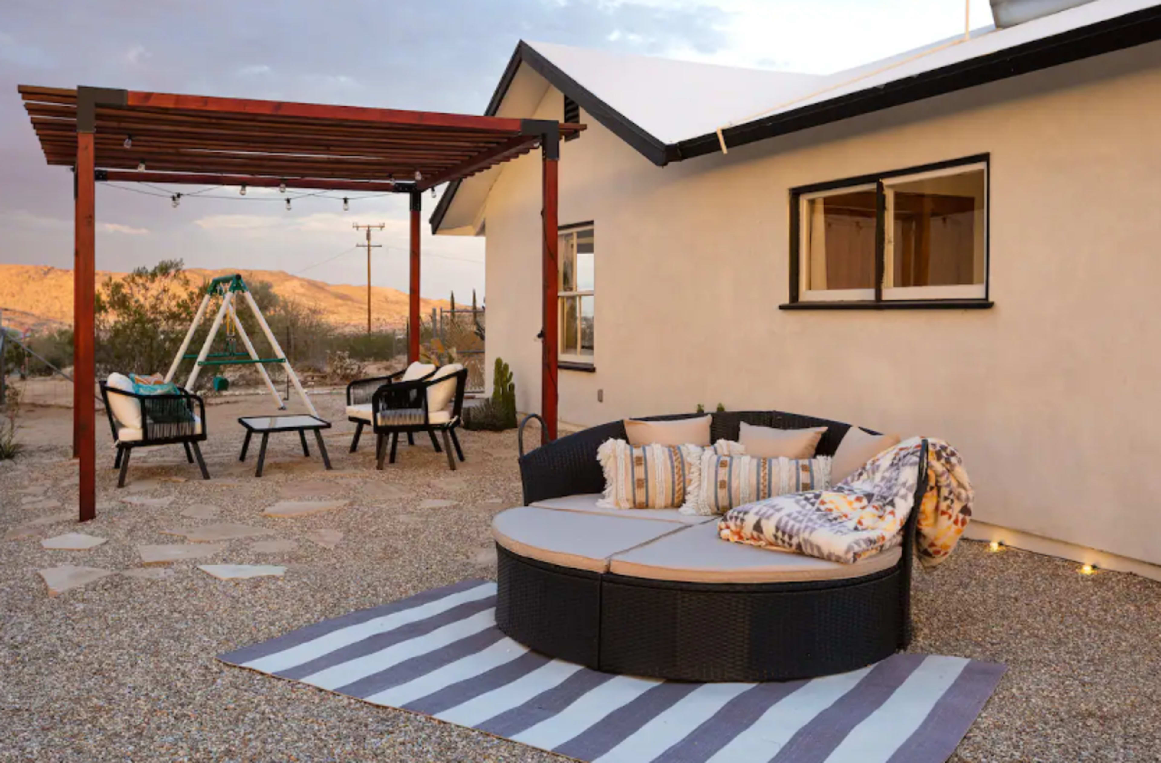 A circular outdoor seating area with cushions is positioned on a gravel patio beside a house and a wooden pergola.