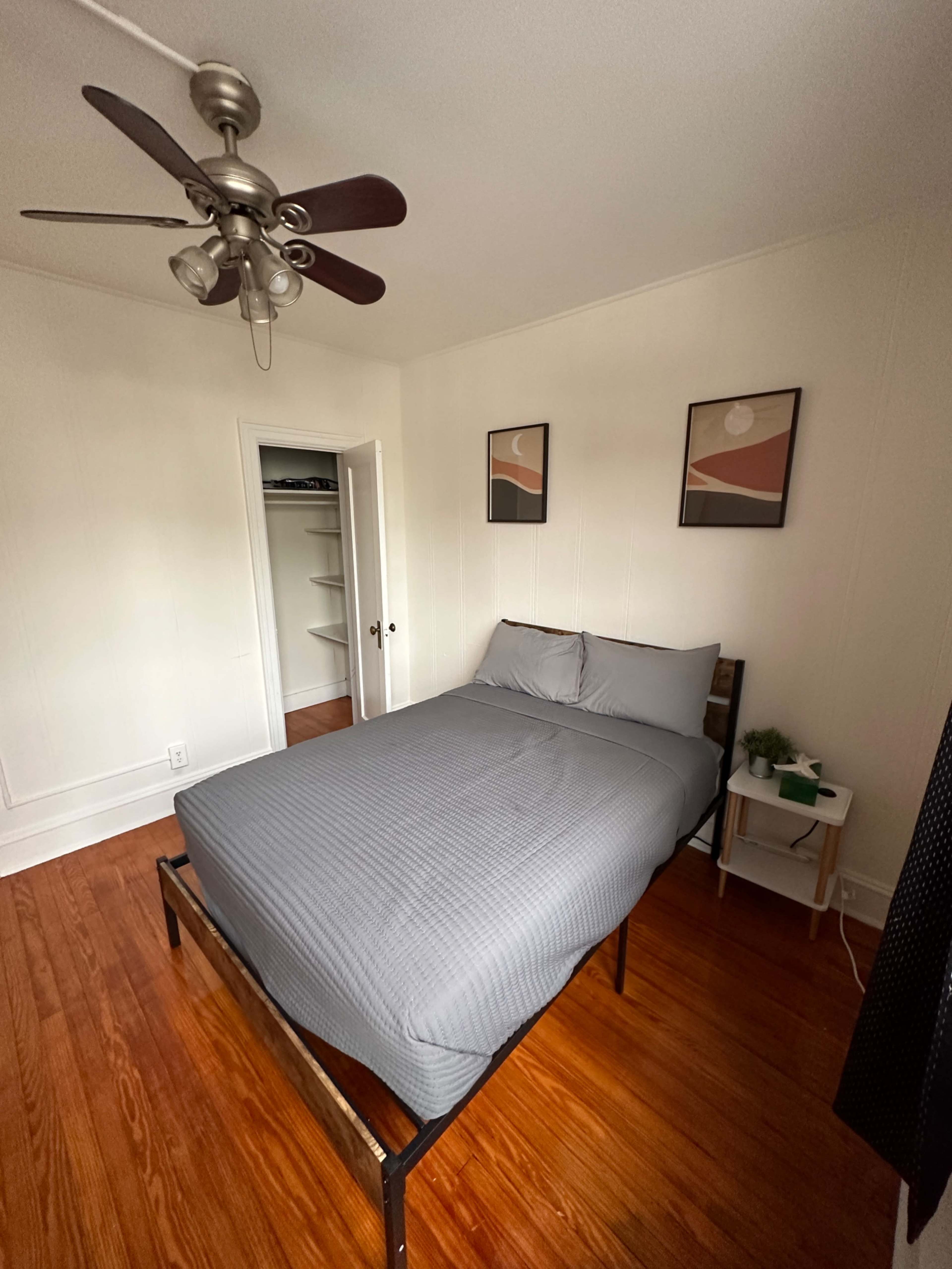 A simple bedroom with a bed, a nightstand, a ceiling fan, and two framed artworks on the walls.