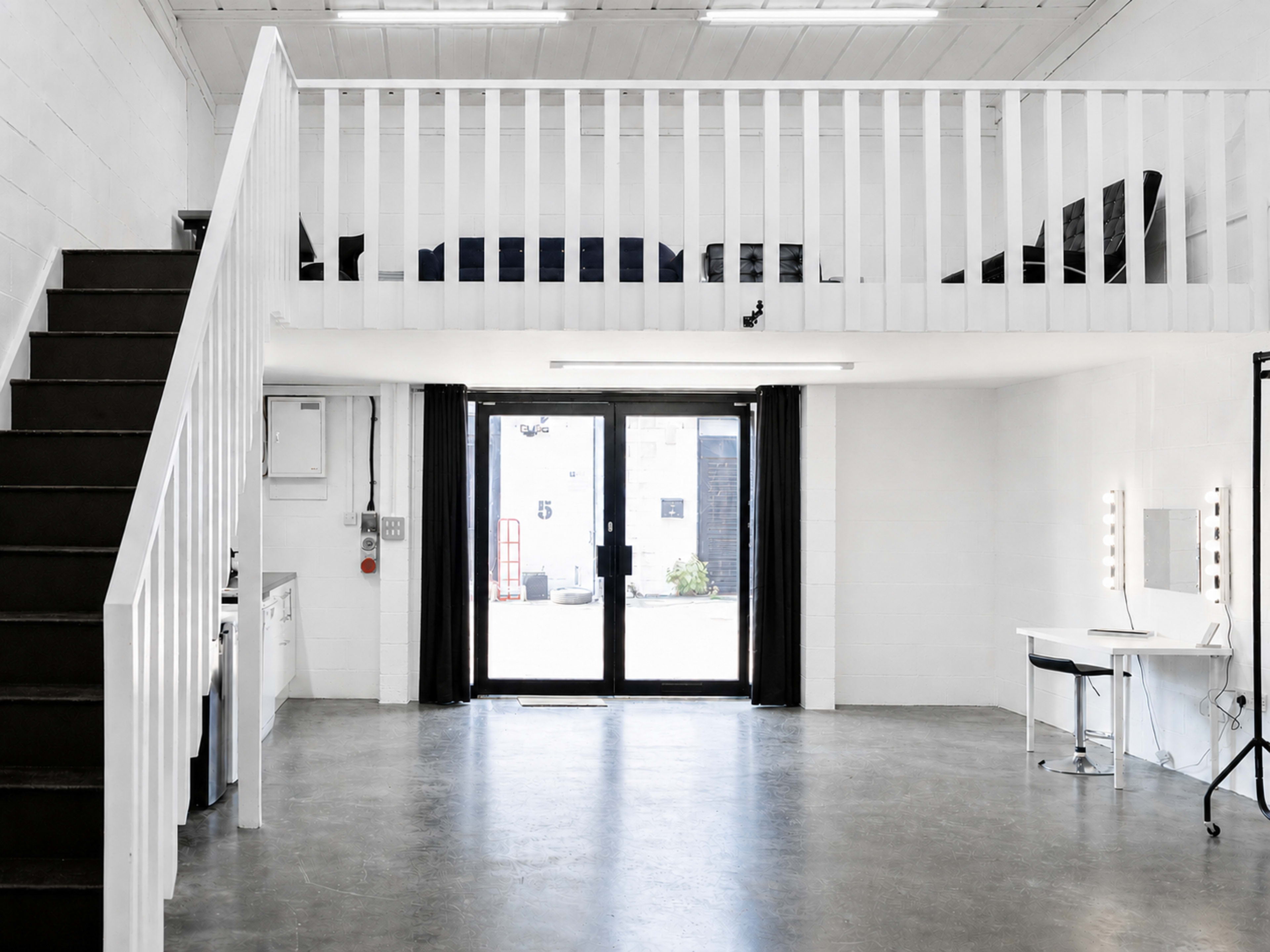 The image shows a spacious interior with a loft area, a staircase on the left, and large glass doors leading to a courtyard, all featuring minimalist decor.
