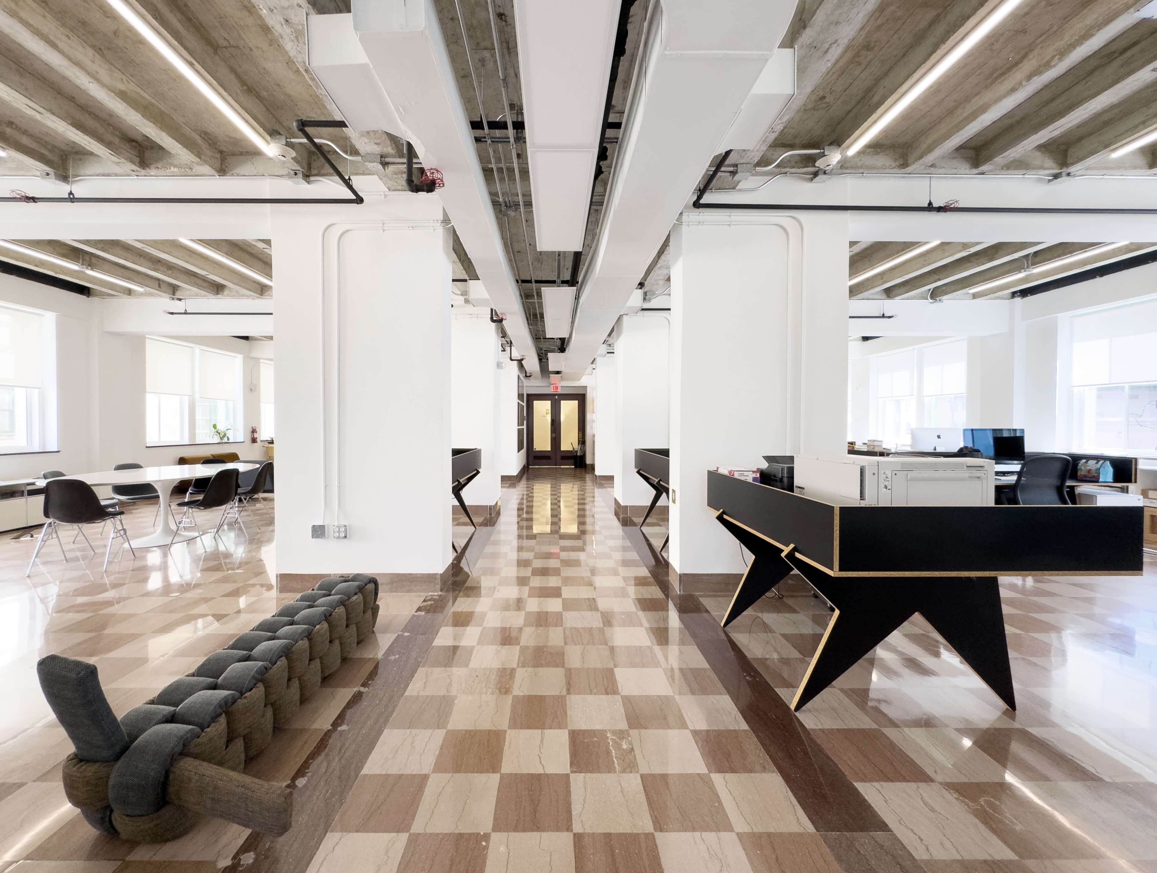 The image shows a spacious modern office interior with a checkered floor, exposed beams, and a mix of seating areas and workstations.