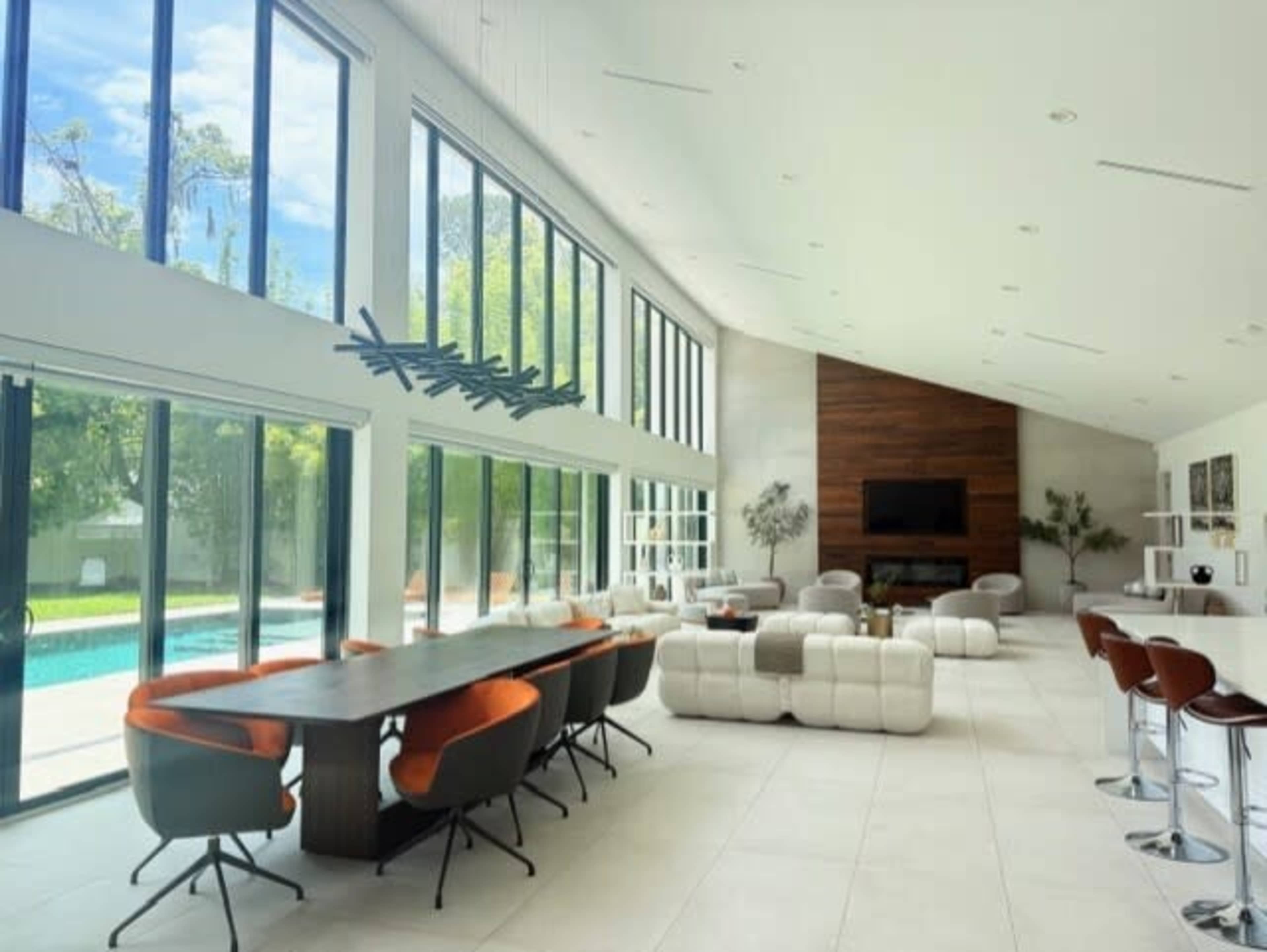 A modern, open-concept living space features large windows, a long dining table with chairs, and a lounge area with white furniture, all overlooking a pool and greenery outside.