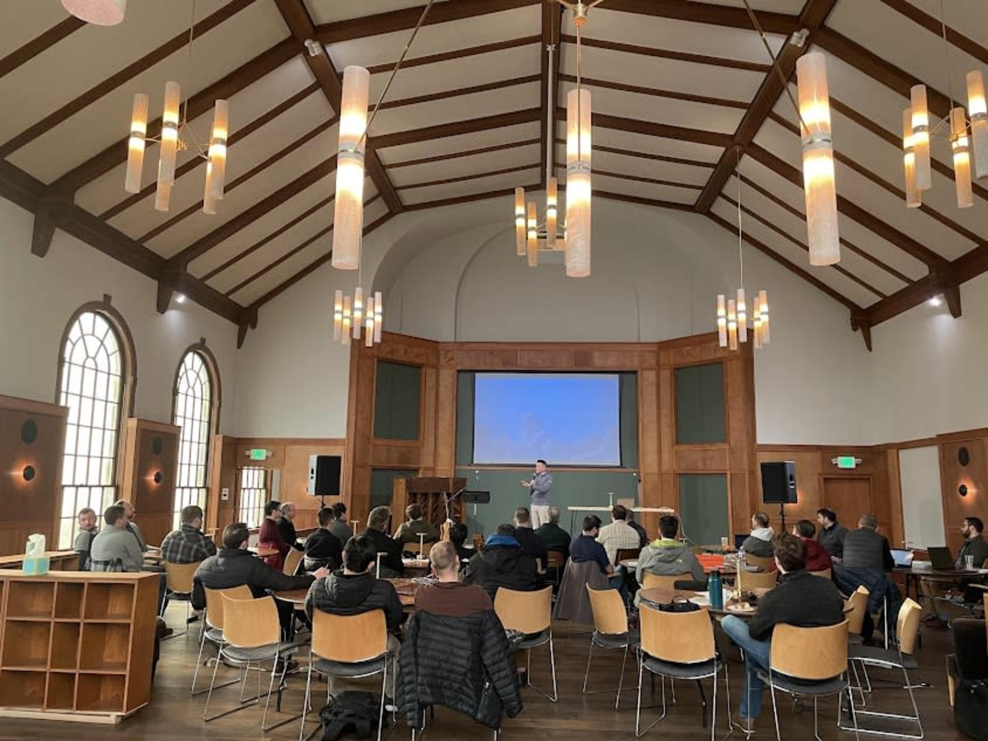 A speaker presents in front of an audience seated in a large, well-lit room with high ceilings and wooden accents.